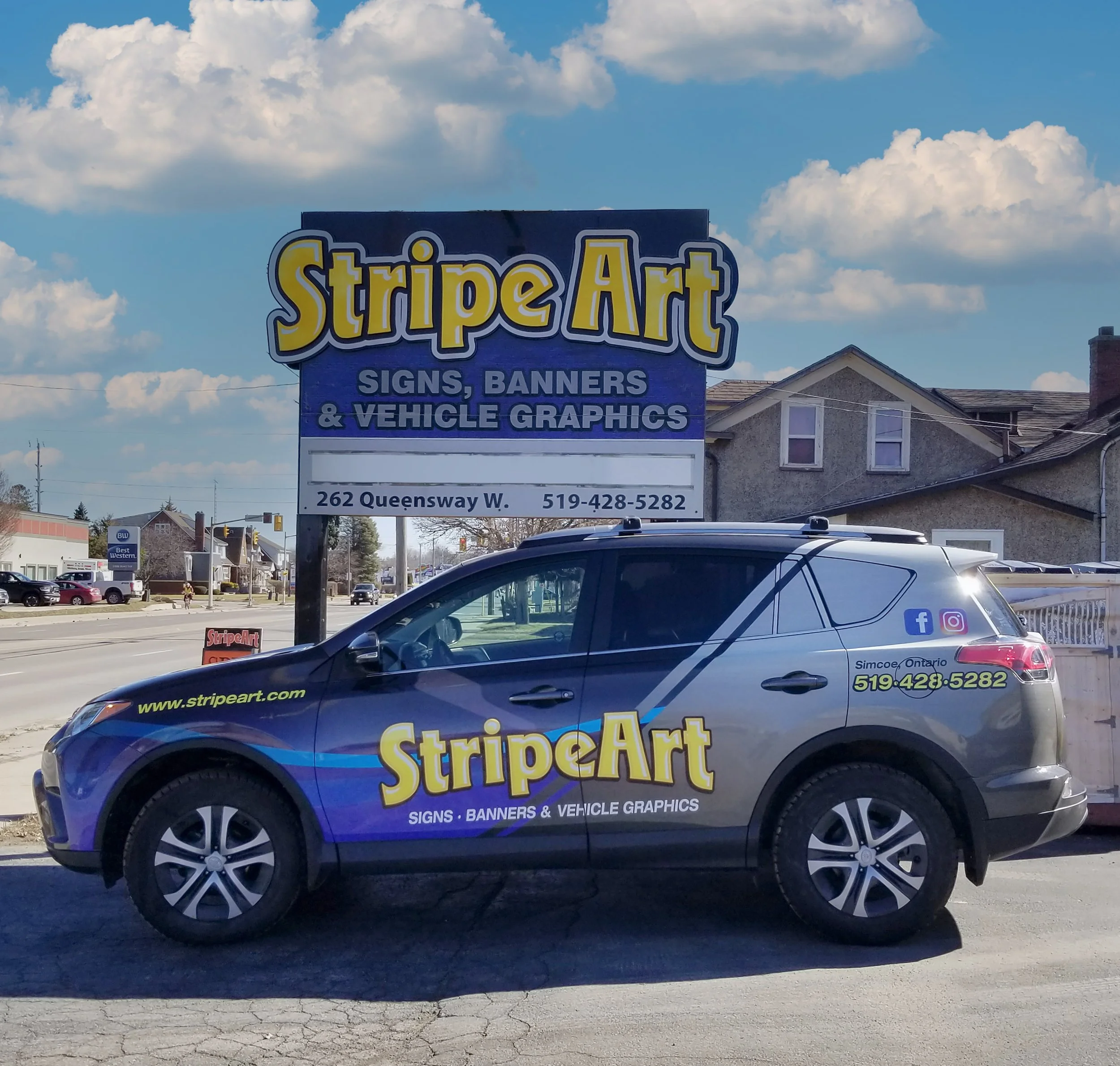 Commercial vehicle with 'StripeArt' branding parked in front of a business sign for StripeArt, a sign, banner, and vehicle graphics company, in a small town with residential houses and a busy street under a partly cloudy sky.