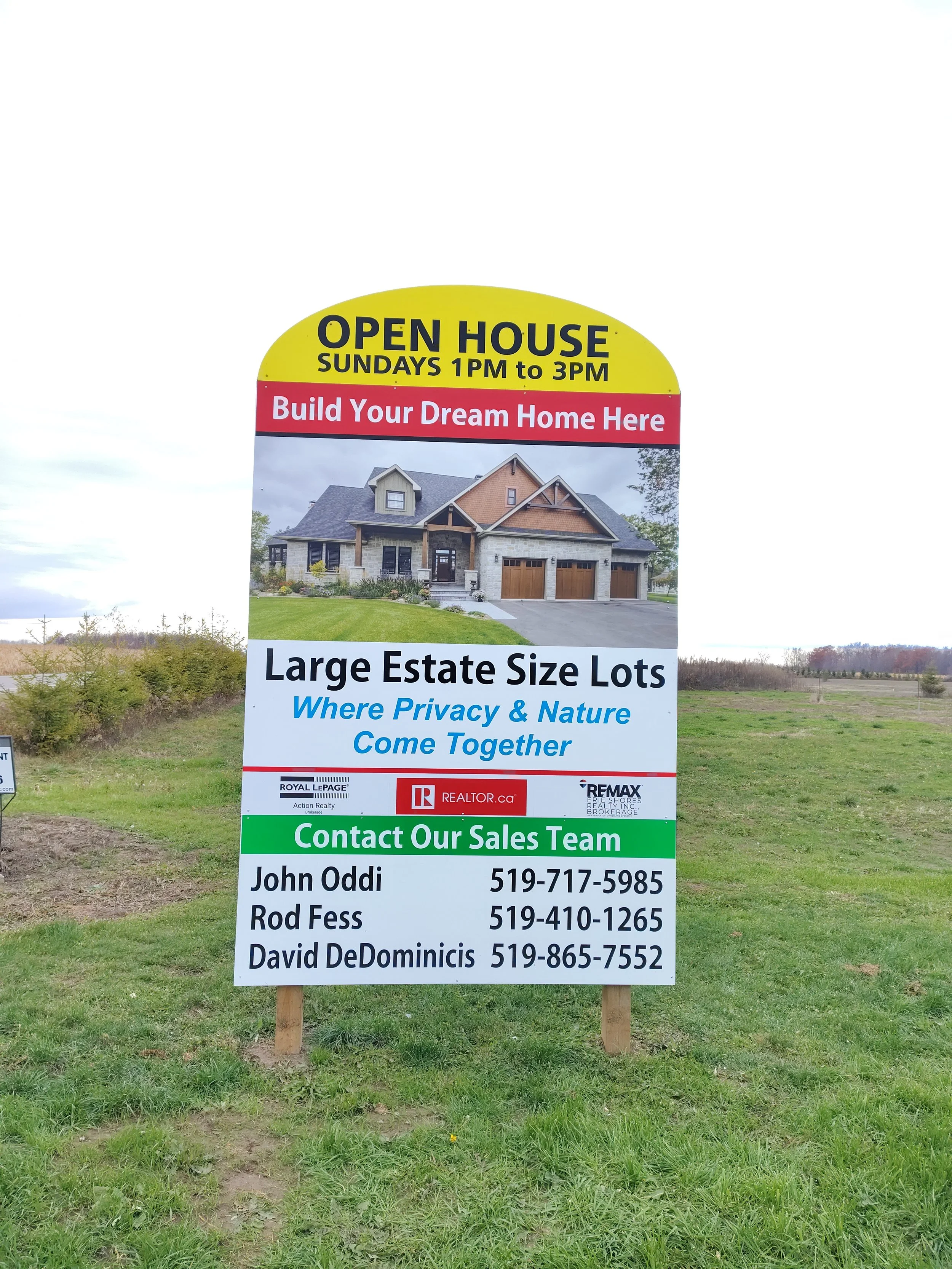 Sign advertising an open house for a new home build, featuring a photograph of a large, modern house with a front lawn. The sign indicates the open house is Sundays from 1 pm to 3 pm, and promotes large estate size lots where privacy and nature come together. It provides contact information for three real estate agents: John Oddi, Rod Fess, and David DeDominicis.