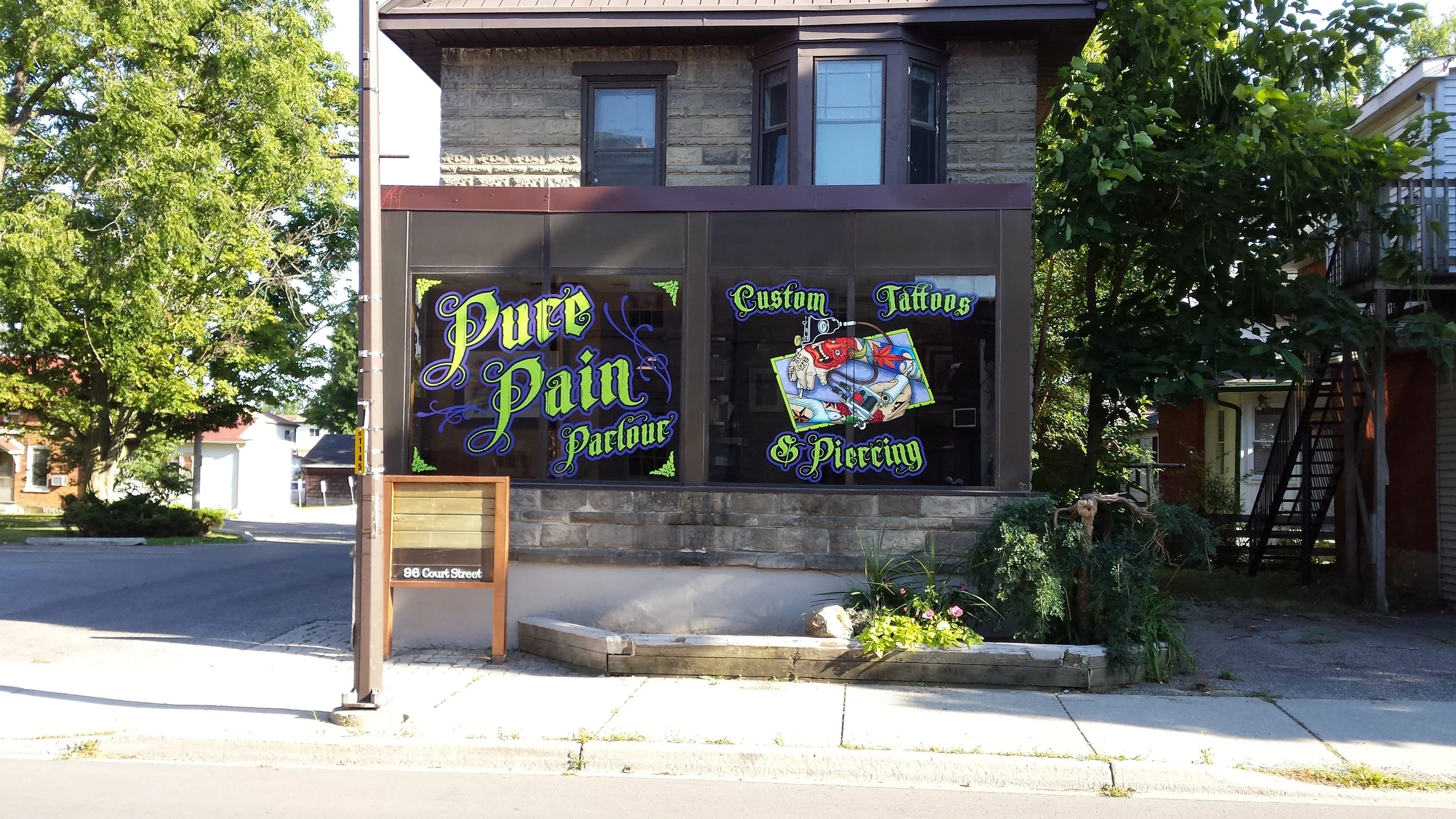 Storefront with sign reading 'Pure Pain Parlour' offering custom tattoos and piercing, located at 96 Court Street, with black window signs in ornate green and purple font, and a small wooden sandwich board in front. There are trees and neighboring buildings around.