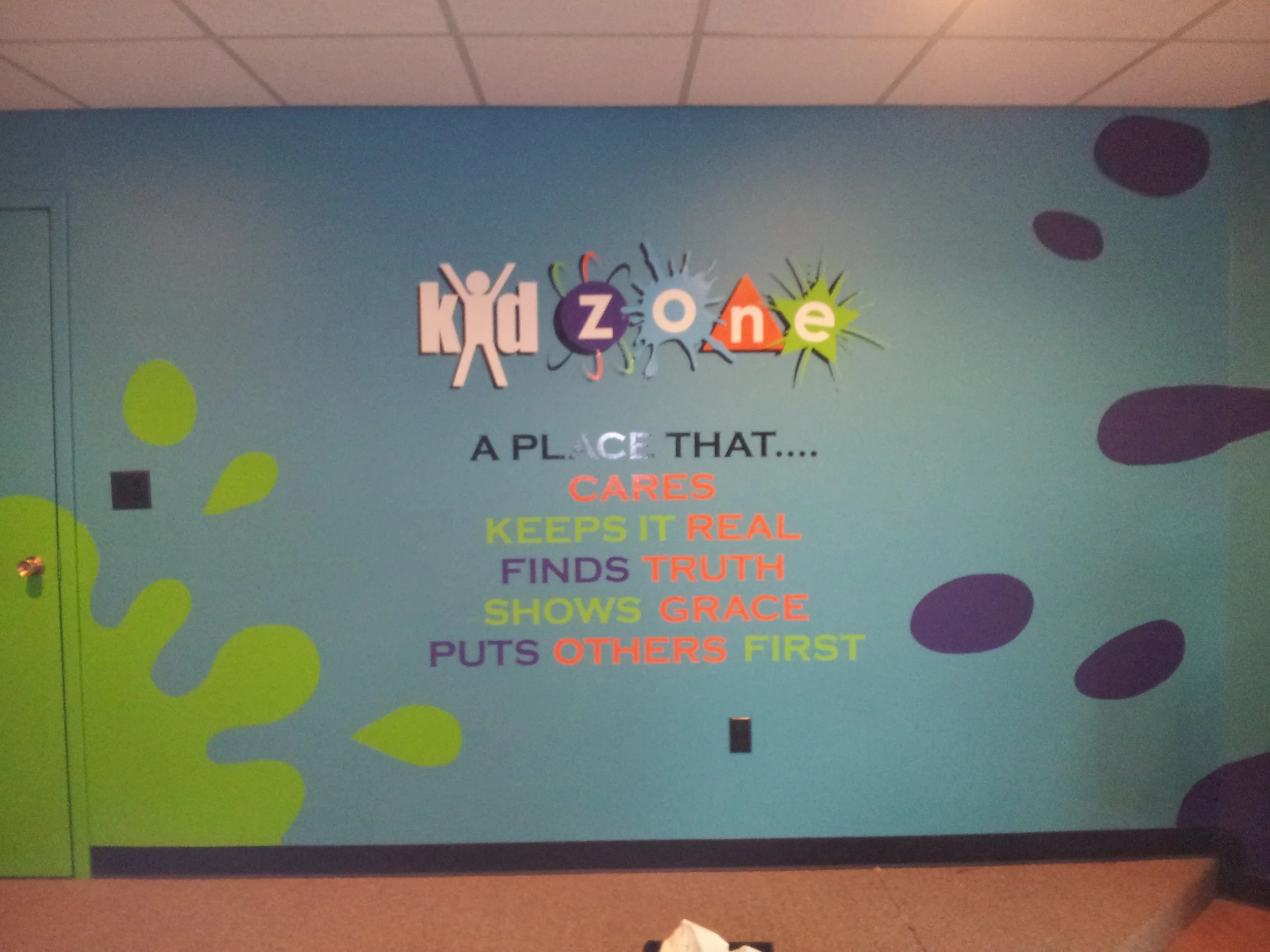 Colorful wall with the word 'kidzone' at the top, decorated with playful shapes and colors. Below it is a list of positive traits written in various colors: 'A place that... cares, keeps it real, finds truth, shows grace, puts others first.'