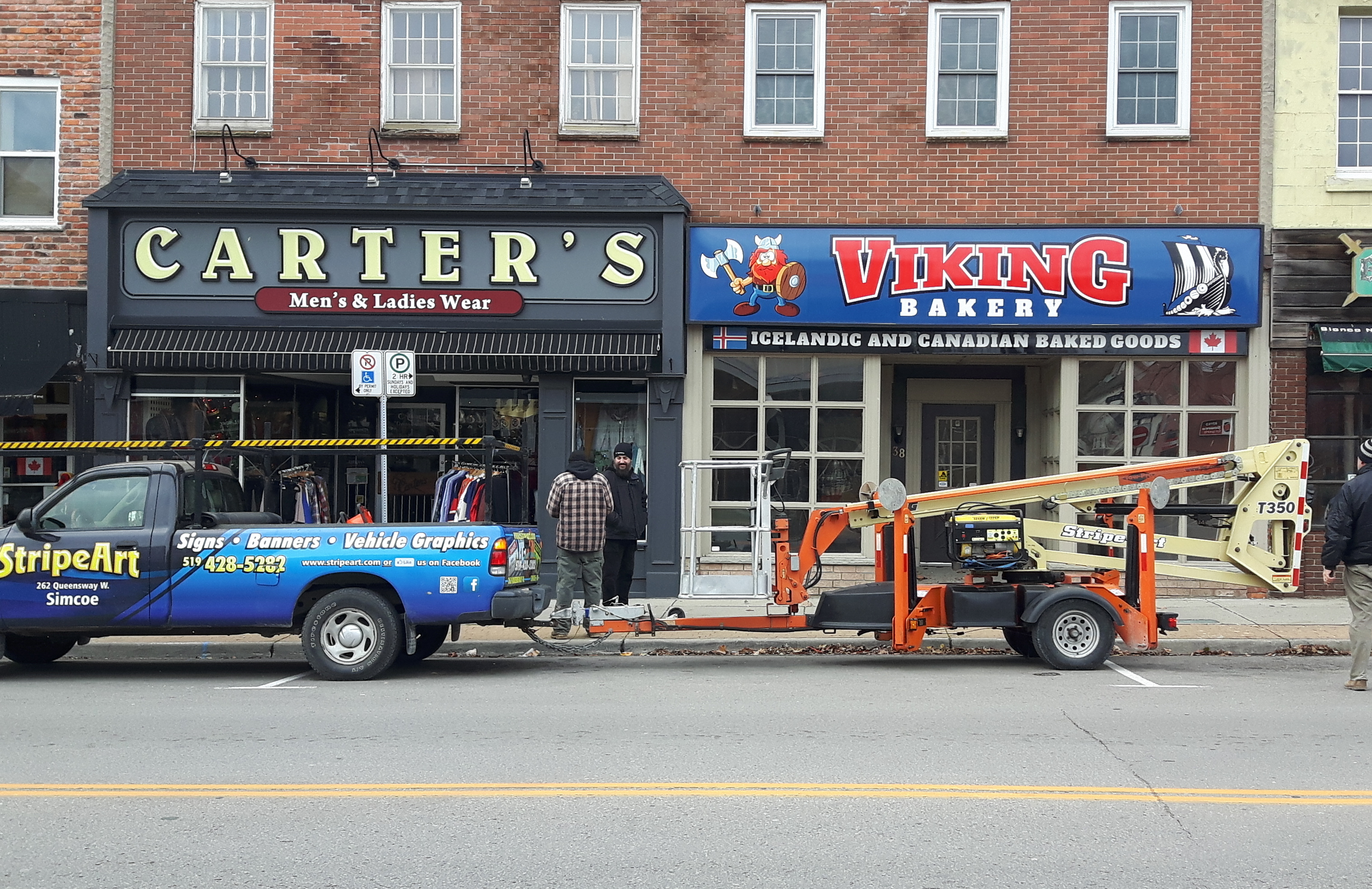 Street view showing a parked sign and vehicle graphics truck in front of a men's and ladies wear store and a bakery with Viking branding, two people talking in front of the stores, and a lifting crane attached to the truck.