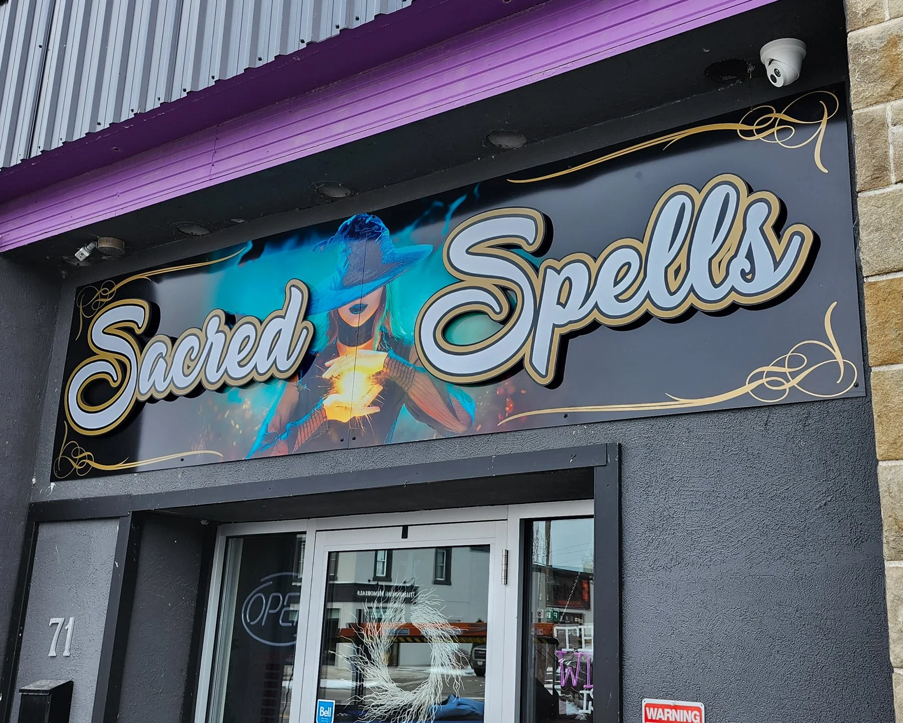 A storefront sign for Sacred Spells with a woman wearing a large hat holding a glowing object, set against a dark background with decorative gold flourishes.