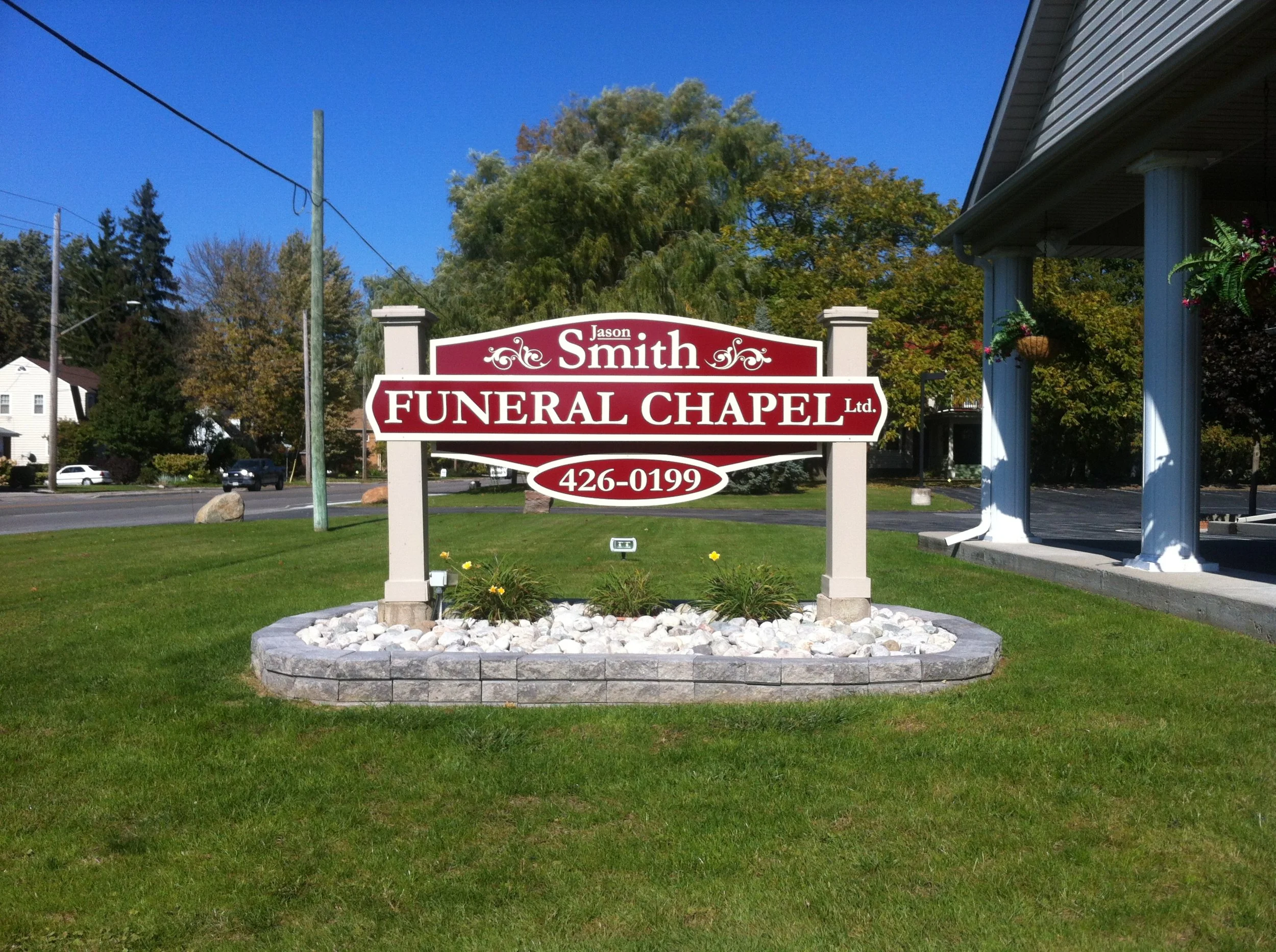 Sign for Jason Smith Funeral Chapel Ltd. with phone number 426-0199, located on a grassy lawn with trees and buildings in the background, under a clear blue sky.