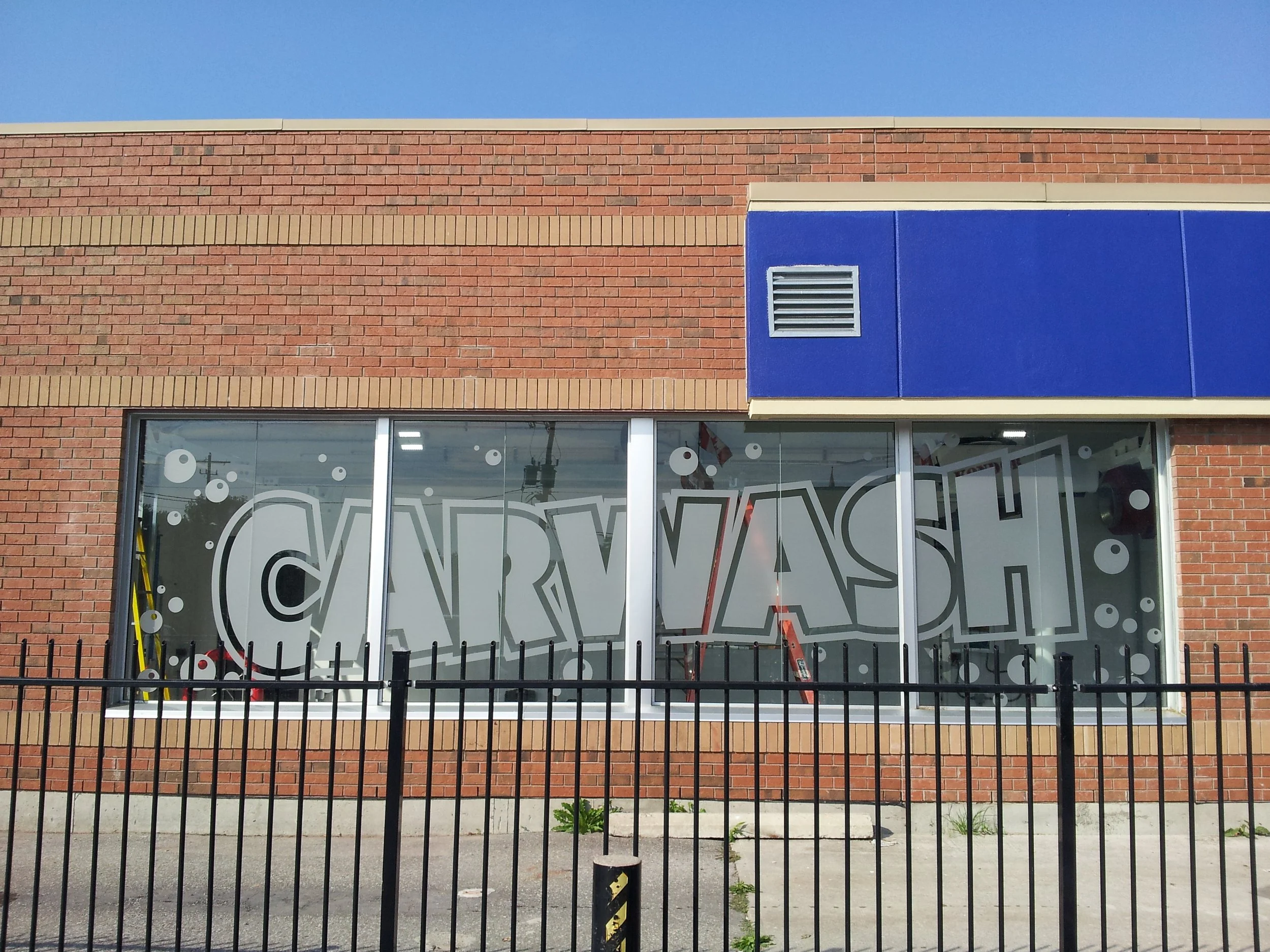 Storefront window with large 'CAR WASH' sign, surrounded by brick wall and a black metal fence in front.