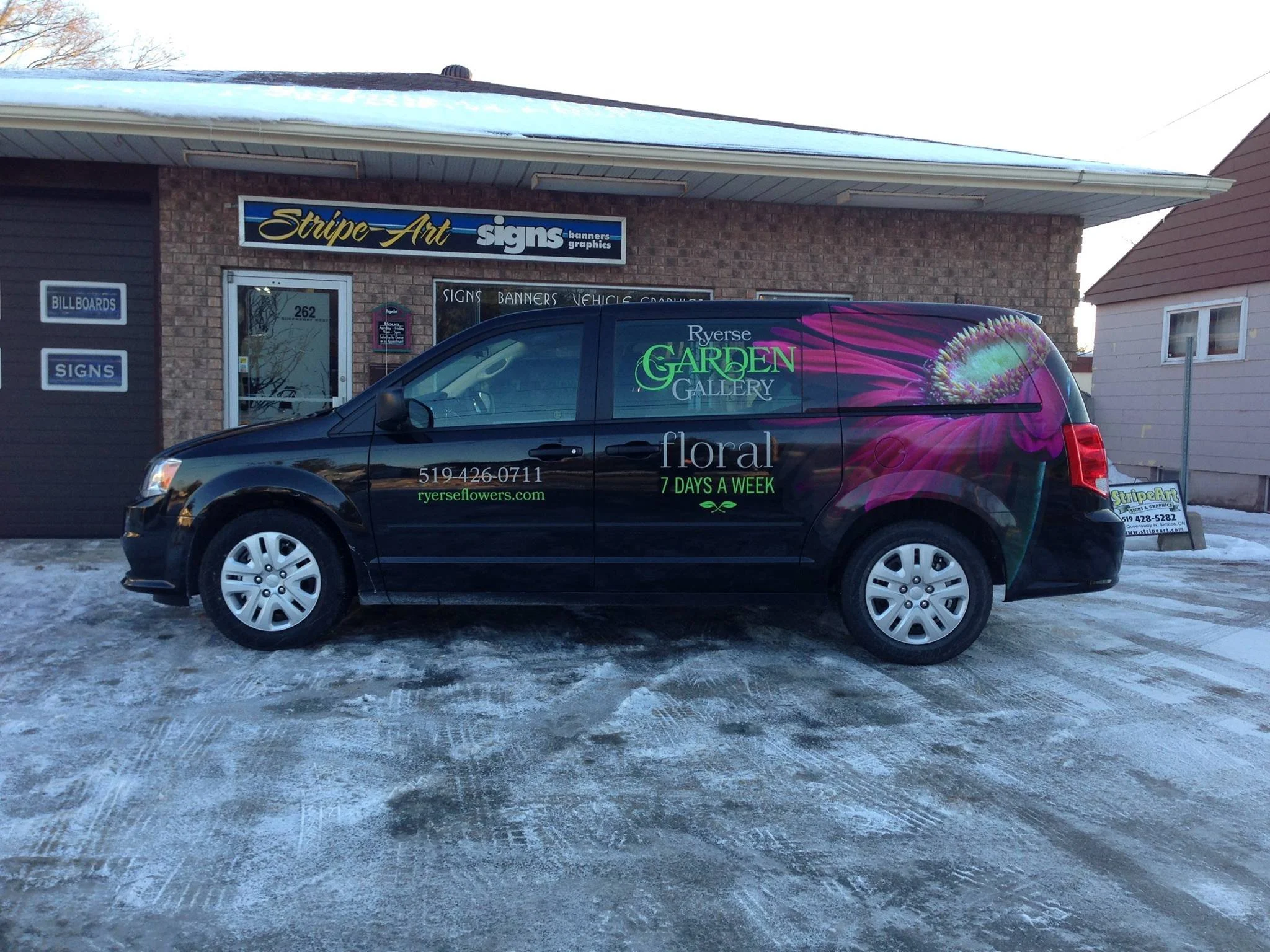 Black van with floral advertisements parked in front of a sign shop on a snowy day.