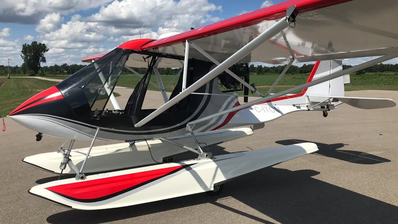 A small white and red hovercraft with a black cockpit sits on an airport tarmac, with green fields and a cloudy sky in the background.