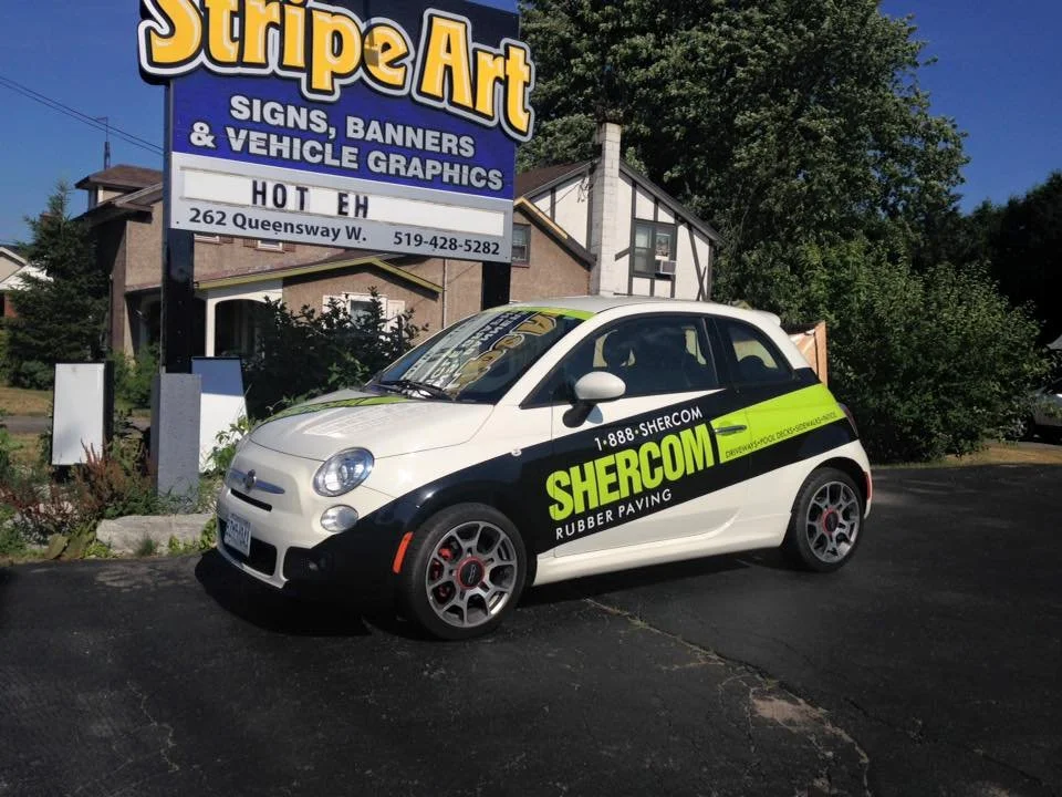 A white and black Shercom delivery car parked in front of a signage for Stripe Art, a business that makes signs, banners, and vehicle graphics, located at 262 Queensway W.