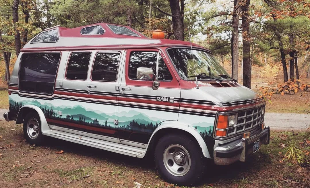A vintage camper van parked in a wooded area with autumn leaves, painted with a mountain landscape mural and a red and white color scheme.