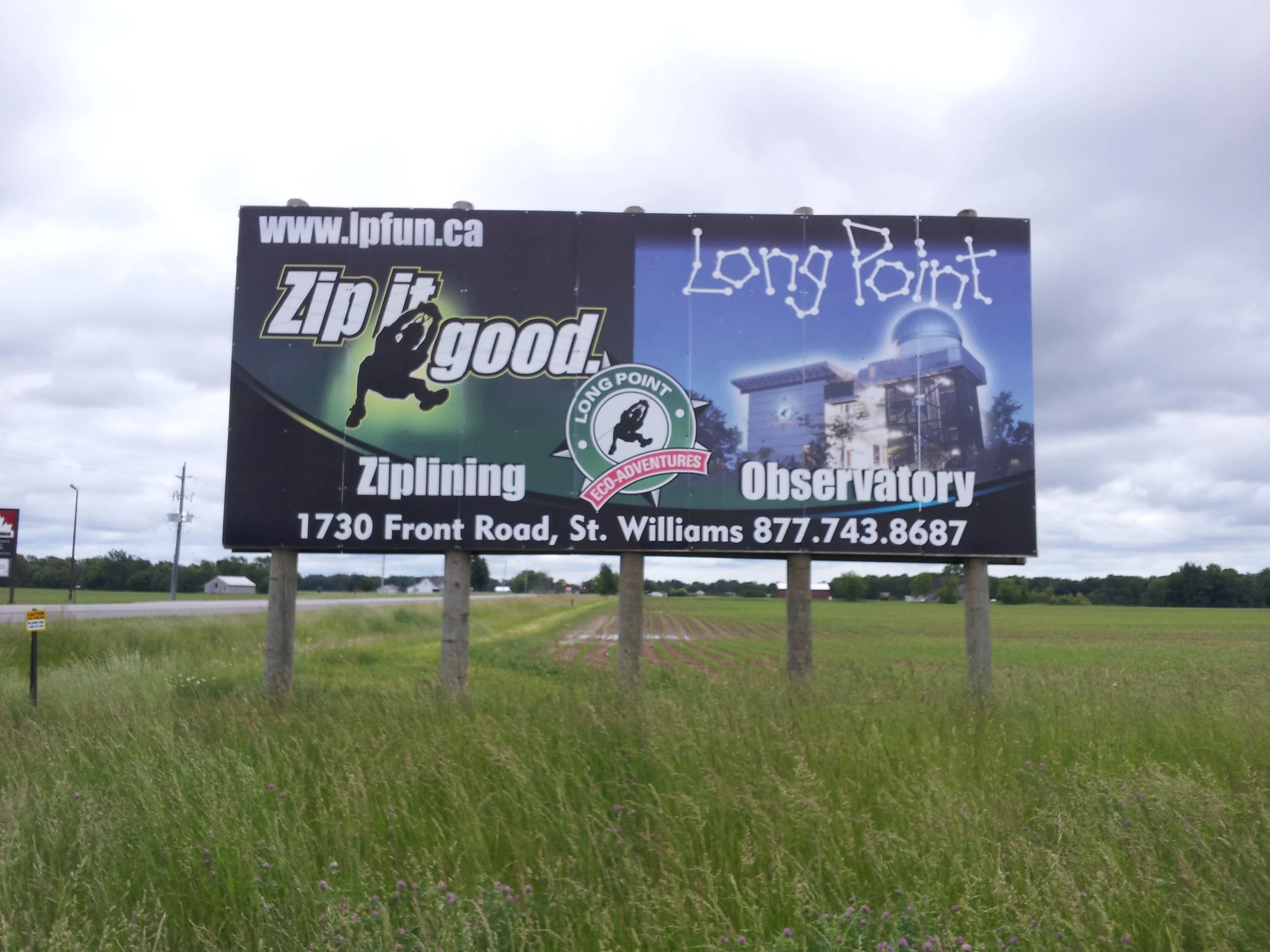 Billboard advertising Long Point Observation Observatory and ziplining, with a picture of a building and constellations, located at 1730 Front Road, St. Williams, with a contact number 877-743-8687, in a rural area with grassy fields and cloudy sky.