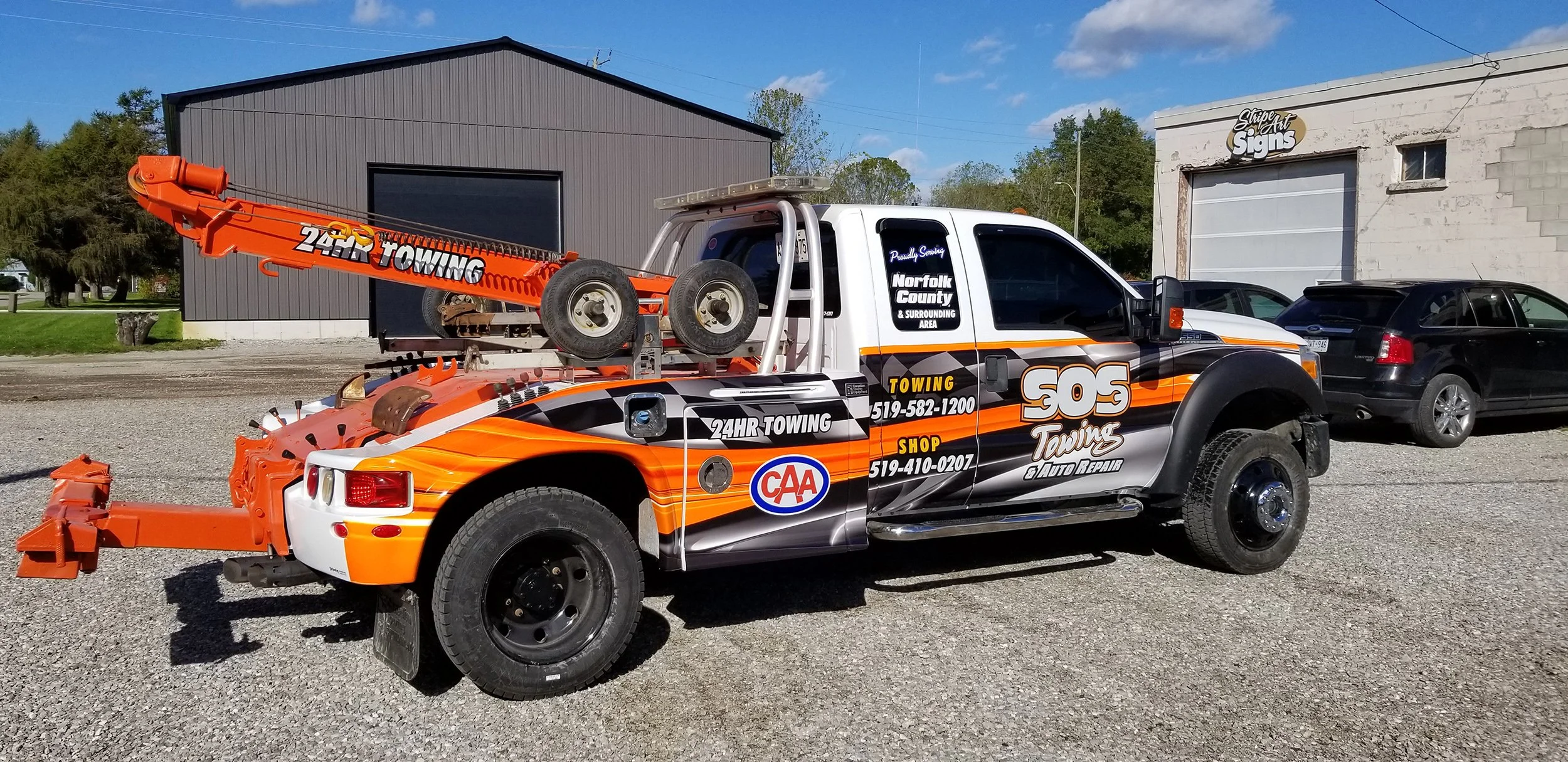 A tow truck with orange and black graphics parked outside a garage, with other vehicles and buildings in the background.
