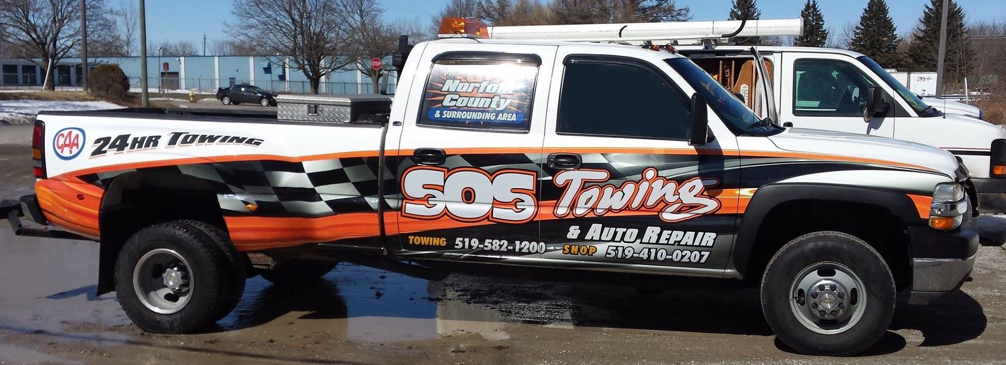 A white tow truck with orange and black racing stripes advertising SOS Towing, with contact information and services, parked on a street during daytime.
