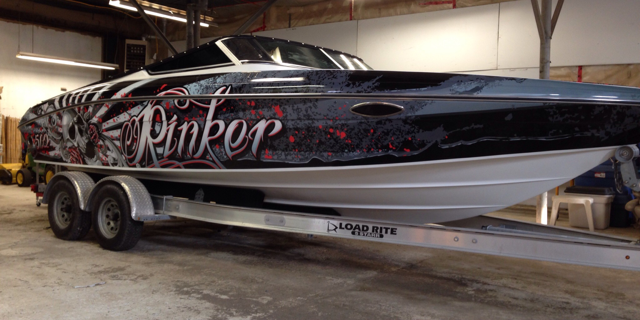 A speedboat with a black, white, and red graffiti-style design and the word 'PinkF' on its side, on a dual-axle trailer inside a wooden building.