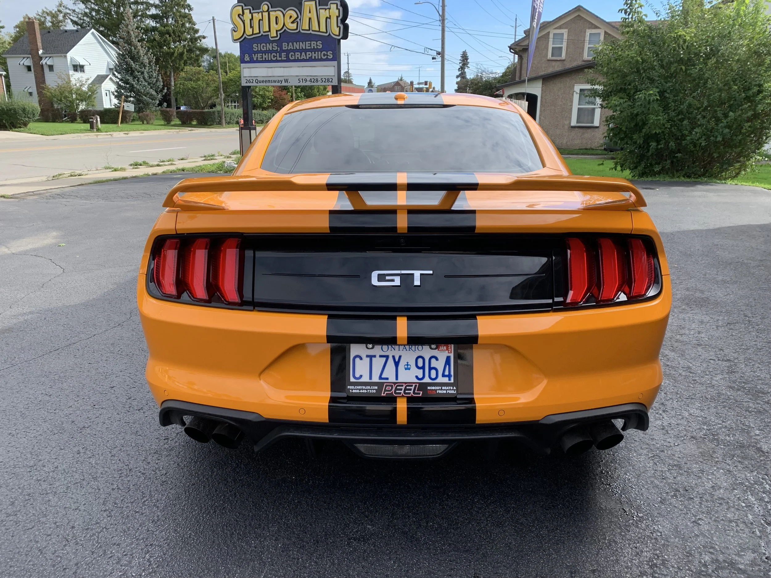 Orange Ford Mustang GT with black racing stripe, parked on asphalt, showing the rear view with taillights, dual exhaust pipes, and Ontario license plate number CTZY 964. In the background, there is a sign for Stripe Art and residential houses.