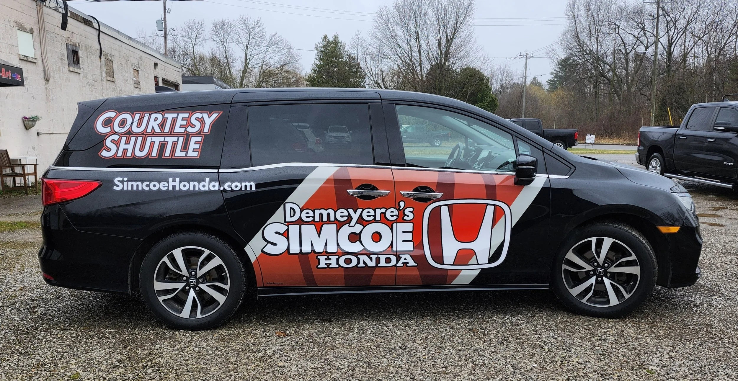 Black Honda minivan with red and white branding for Demerey's Simcoe Honda, displaying website and shuttle service information on the side, parked on gravel with other vehicles and a building in the background.