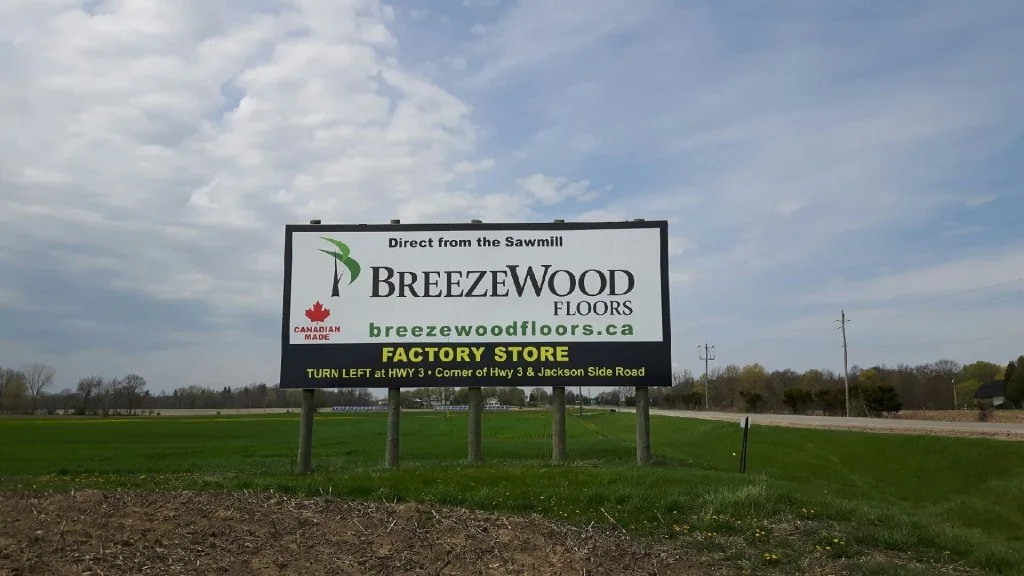 A large roadside billboard advertising BreezeWood Floors, a Canadian-made flooring company, with directions to their factory store, located at the corner of HWY 3 and Jackson Side Road.