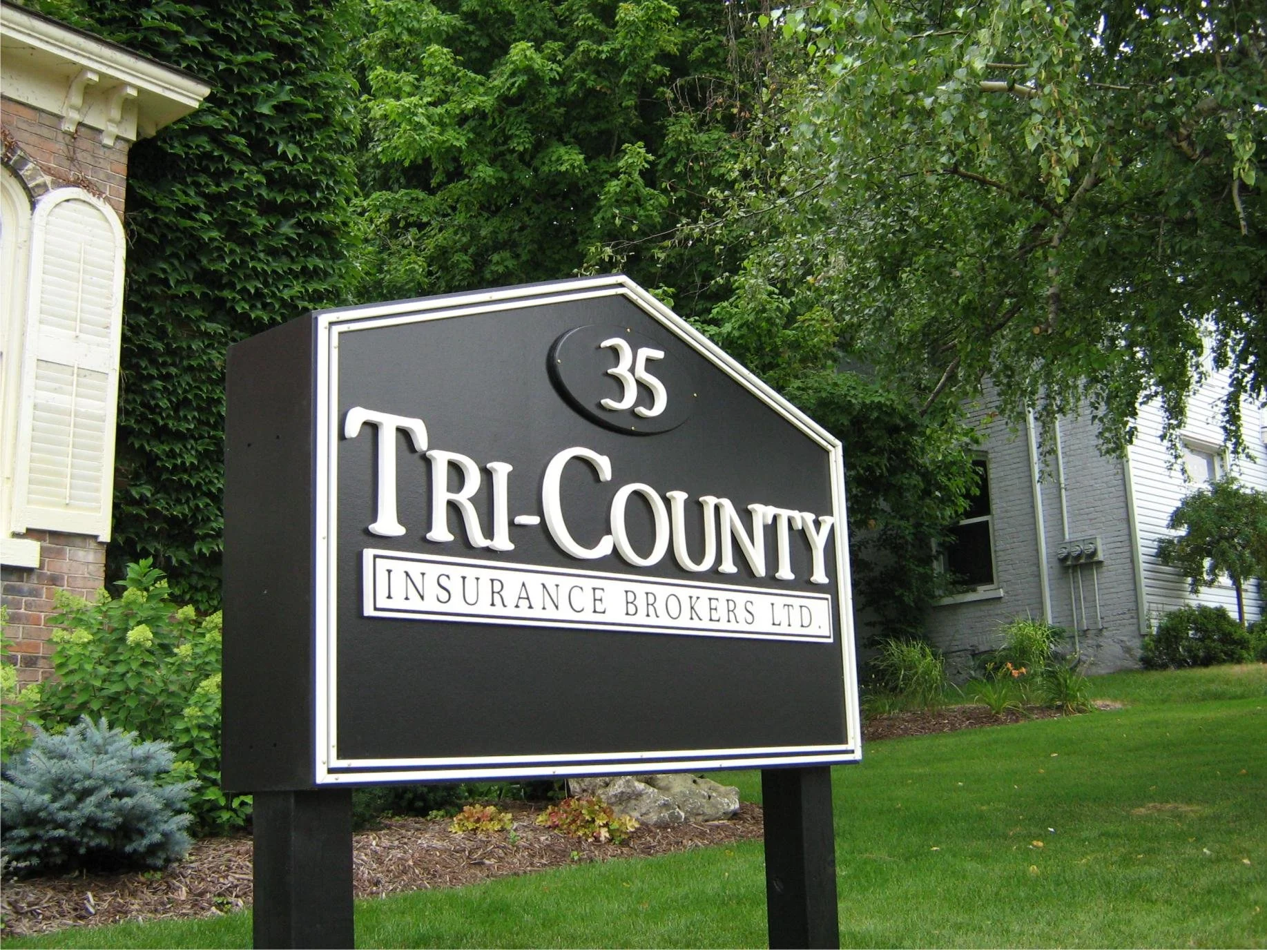 A black sign in front of a residential building reads '35 TRI-COUNTY INSURANCE BROKERS LTD.' with a white border and text. Green trees and shrubs are in the background.