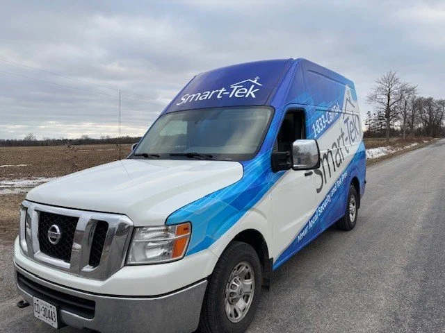 White and blue Smart-Tek service van parked on the side of a rural road with a cloudy sky and barren trees in the background.