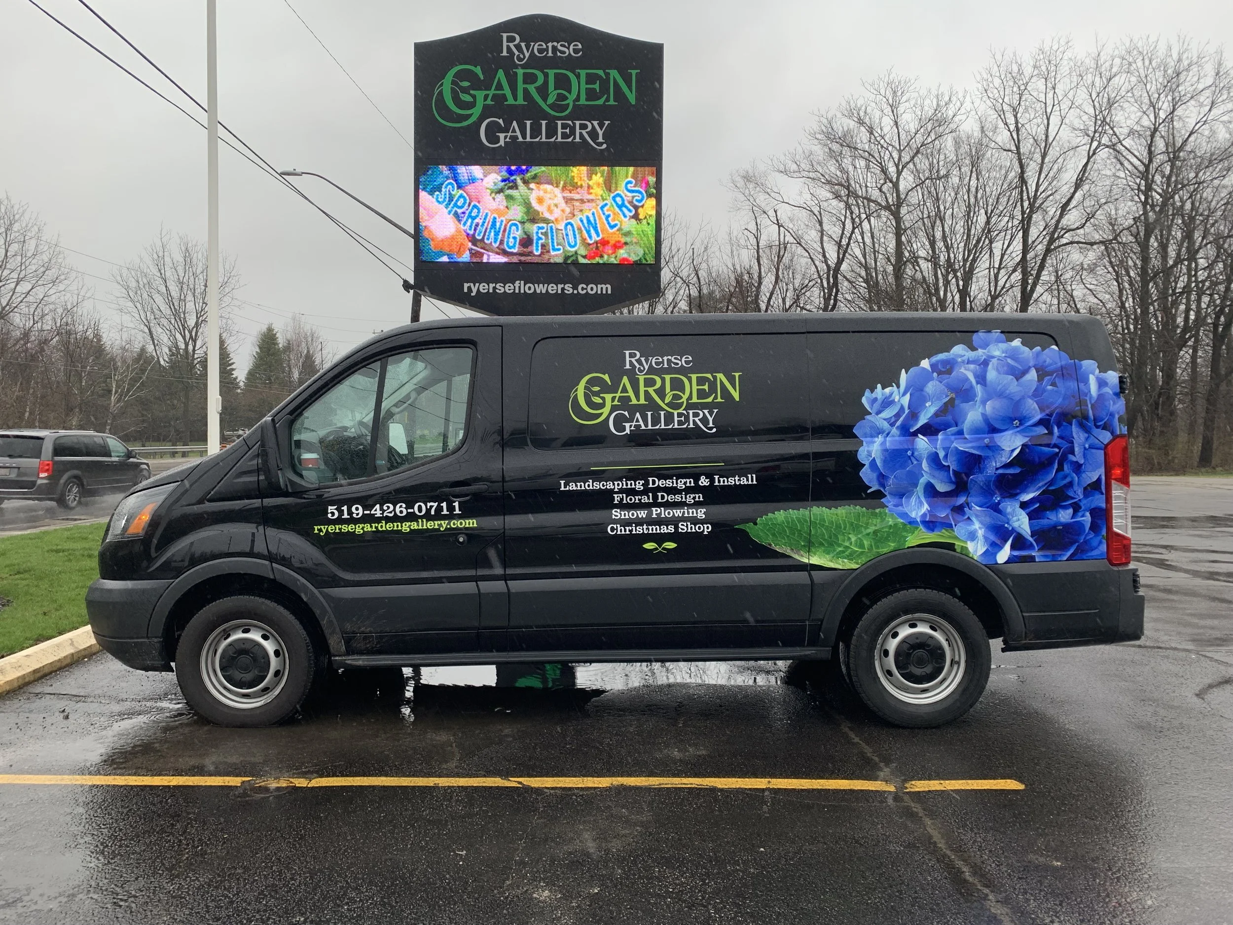 Black van with colorful floral graphics and the text 'Ryerse Garden Gallery' on the side, parked in a lot on a rainy day. Billboard above advertising 'Spring Flowers' and 'Ryerse Flowers' with a website URL.