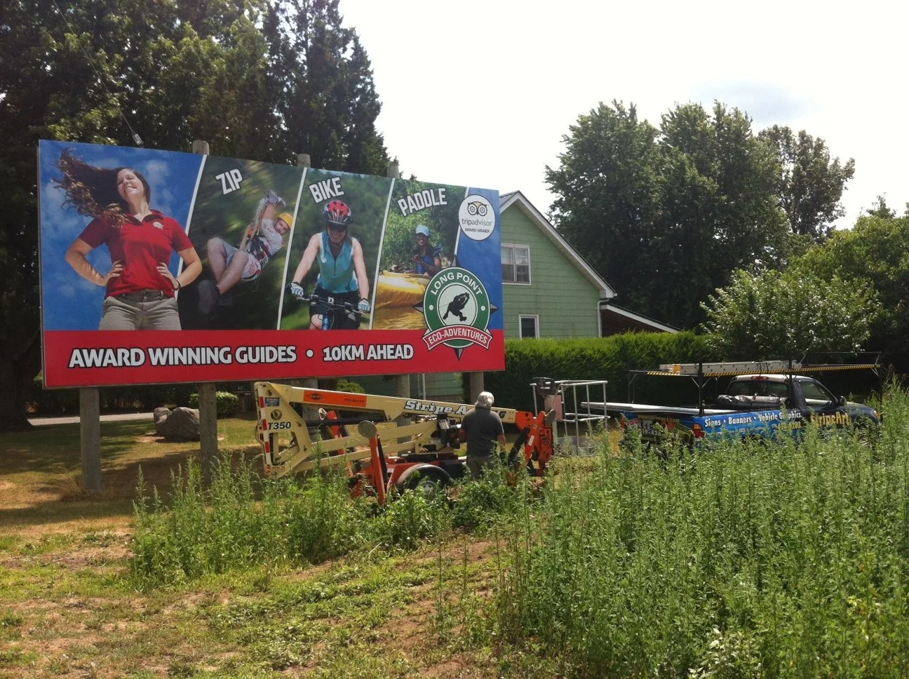 A large outdoor billboard advertising outdoor adventure activities like zip lining, biking, and paddling, featuring images of women engaging in these activities, with a house and trees in the background. There are workmen and equipment near the billboard, and tall grass in the foreground.