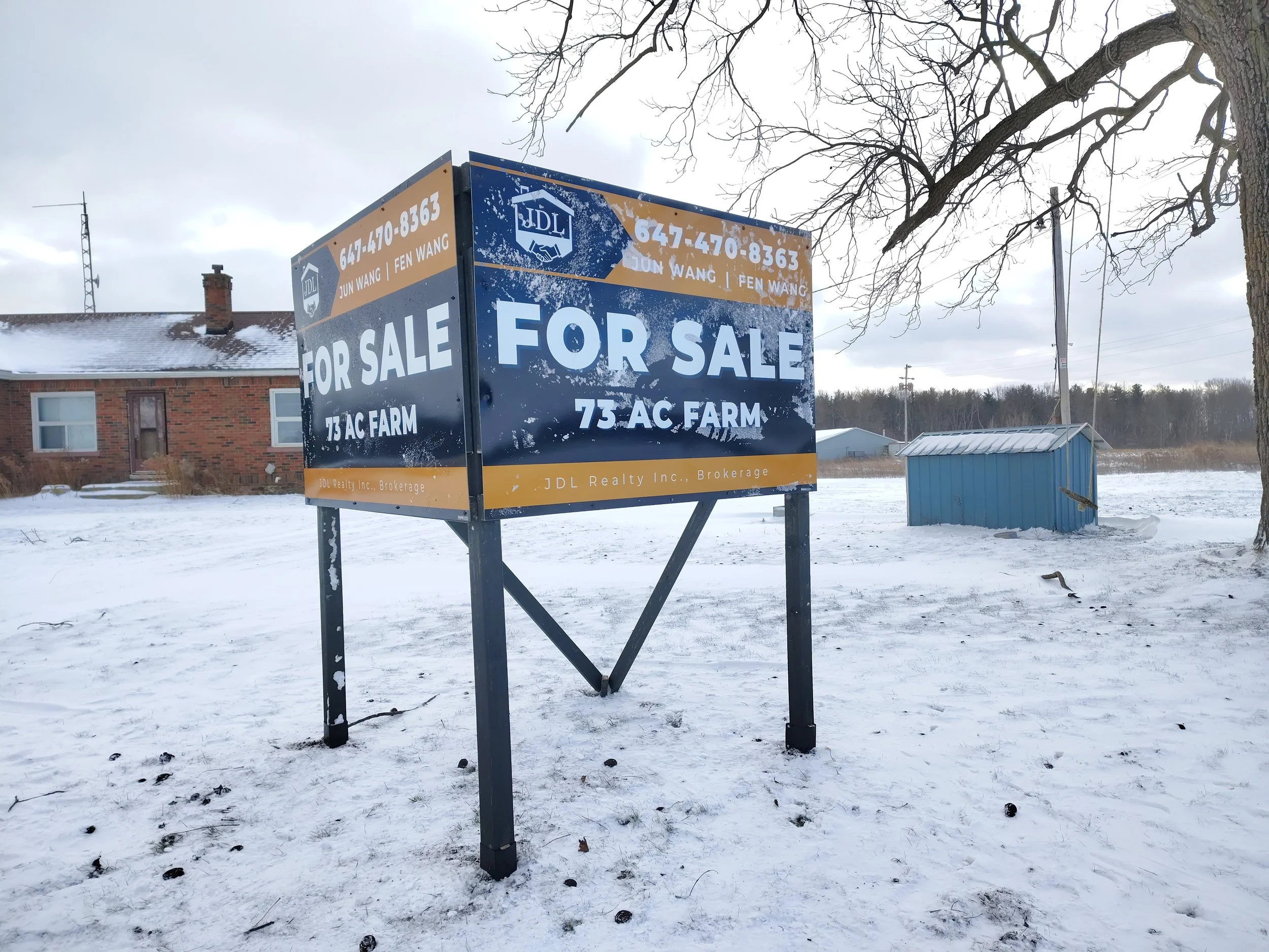 Large blue and white sign with yellow accents advertising a 75-acre farm for sale, placed outdoors in a snowy landscape next to a tree with leafless branches. In the background, there is a brick house with a chimney and snow on its roof, and a small blue building.