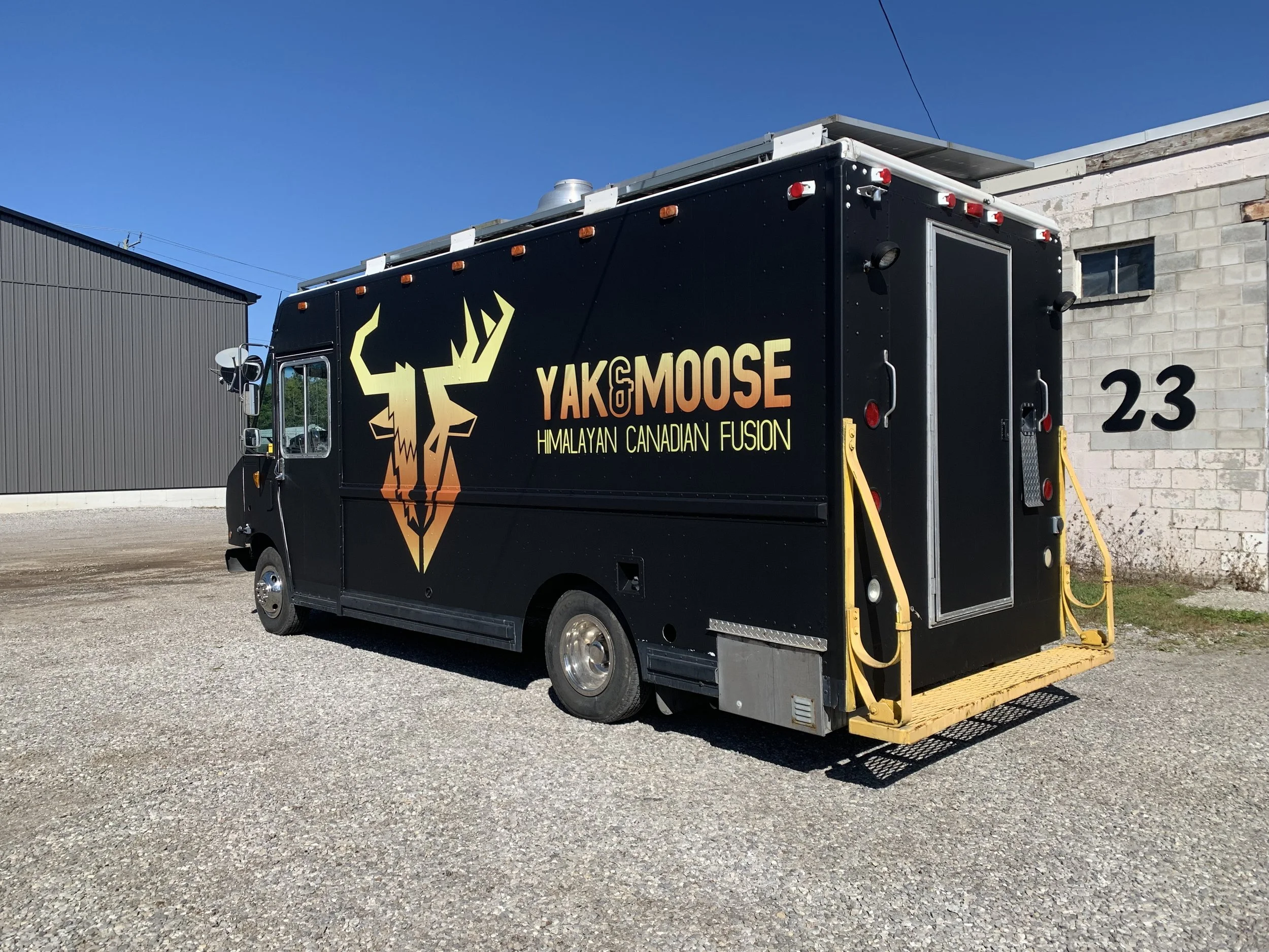 Black food truck with yellow-orange logo of a deer head and text 'YAK MOOSE Himalayan Canadian Fusion' parked outside on gravel ground with warehouse buildings in the background.