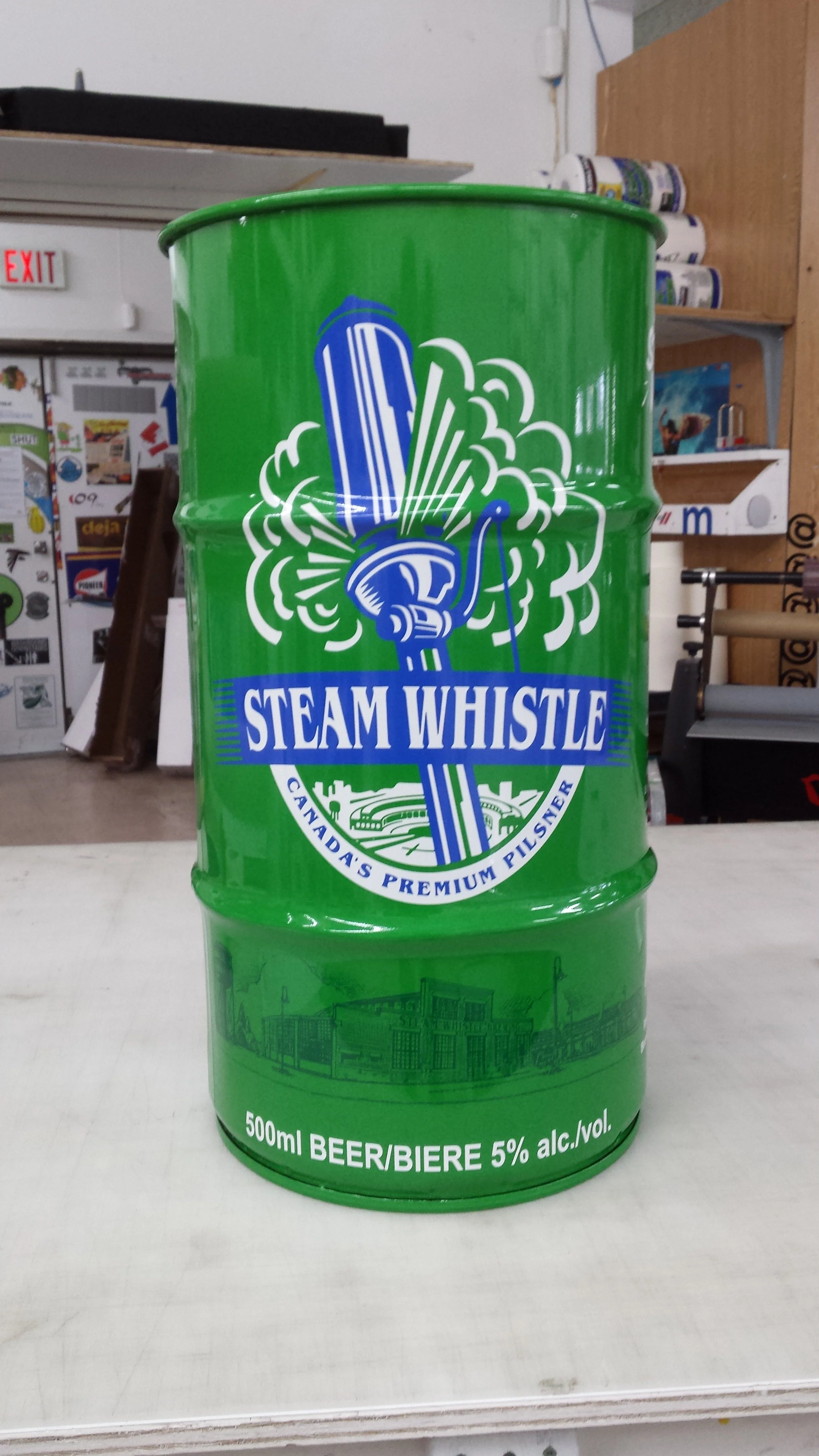 A green metal drum with the label 'Steam Whistle, Canada's premium pilsner,' featuring a graphic of a whistle emitting steam, and text indicating 500ml of beer with 5% alcohol content.