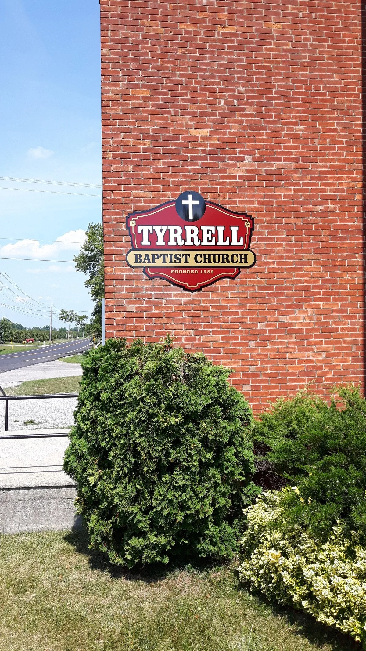 Sign on a red brick wall reading 'Tyrrell Baptist Church, Founded 1859' with a cross symbol above it. Green bushes and plants are in the foreground, and a clear blue sky with a few clouds and a road with utility poles is visible in the background.