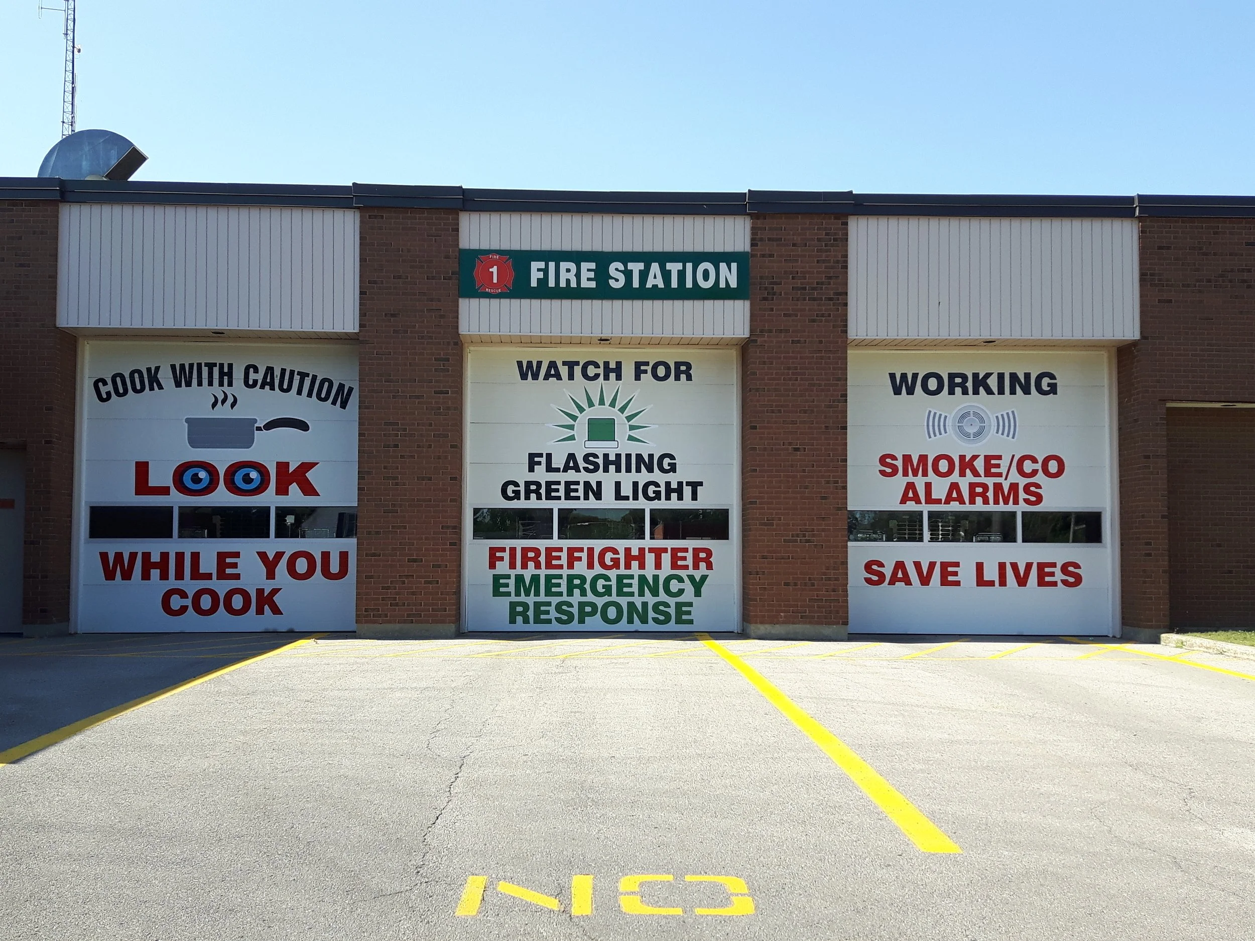 Exterior of a fire station with three garage doors. The left door has a sign that says 'Cook with caution while you cook' and a graphic of a pan, the middle door has a sign that says 'Watch for flashing green light firefighter emergency response' with a graphic of a flashing green light, and the right door has a sign that says 'Working smoke/co alarms save lives' with a graphic of a smoke alarm. The fire station has a brick facade and a green sign above the doors labeled 'Fire Station'.