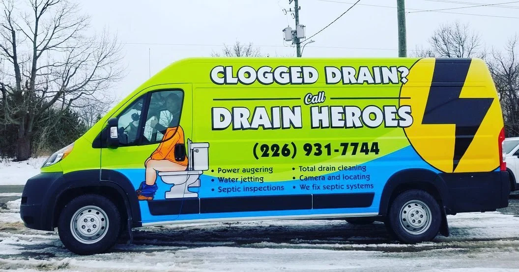 A brightly colored service van advertising clogged drain and drain cleaning services, with a cartoon illustration of a child on a toilet and a large lightning bolt symbol.