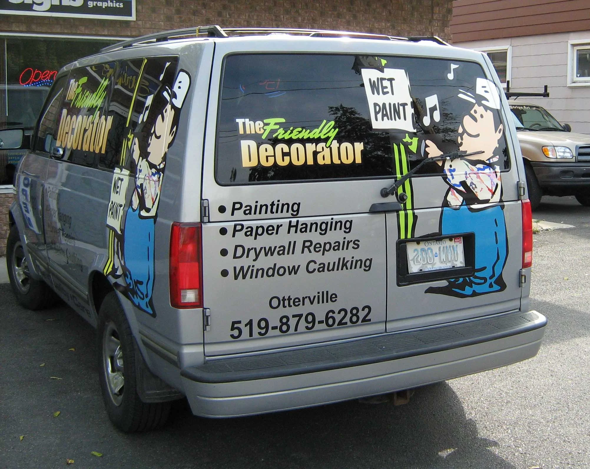 Silver van with colorful cartoon graphics advertising The Friendly Decorator, a painting and drywall repair service, parked on a street.
