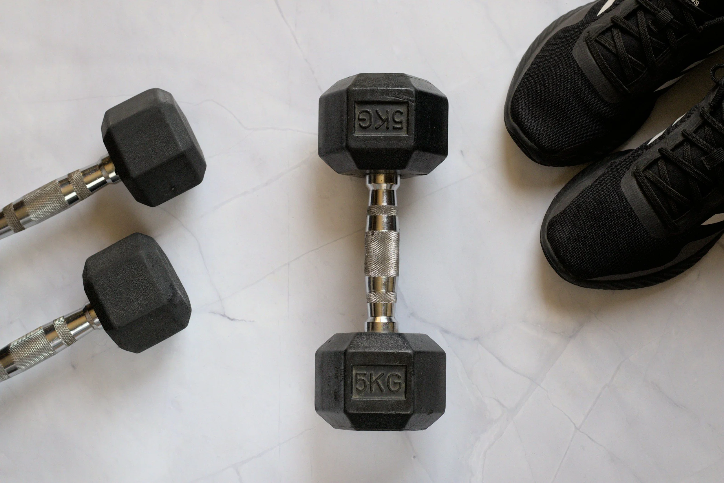 A pair of black dumbbells labeled '5KG', another pair of black dumbbells, and a pair of black athletic shoes on a white surface.