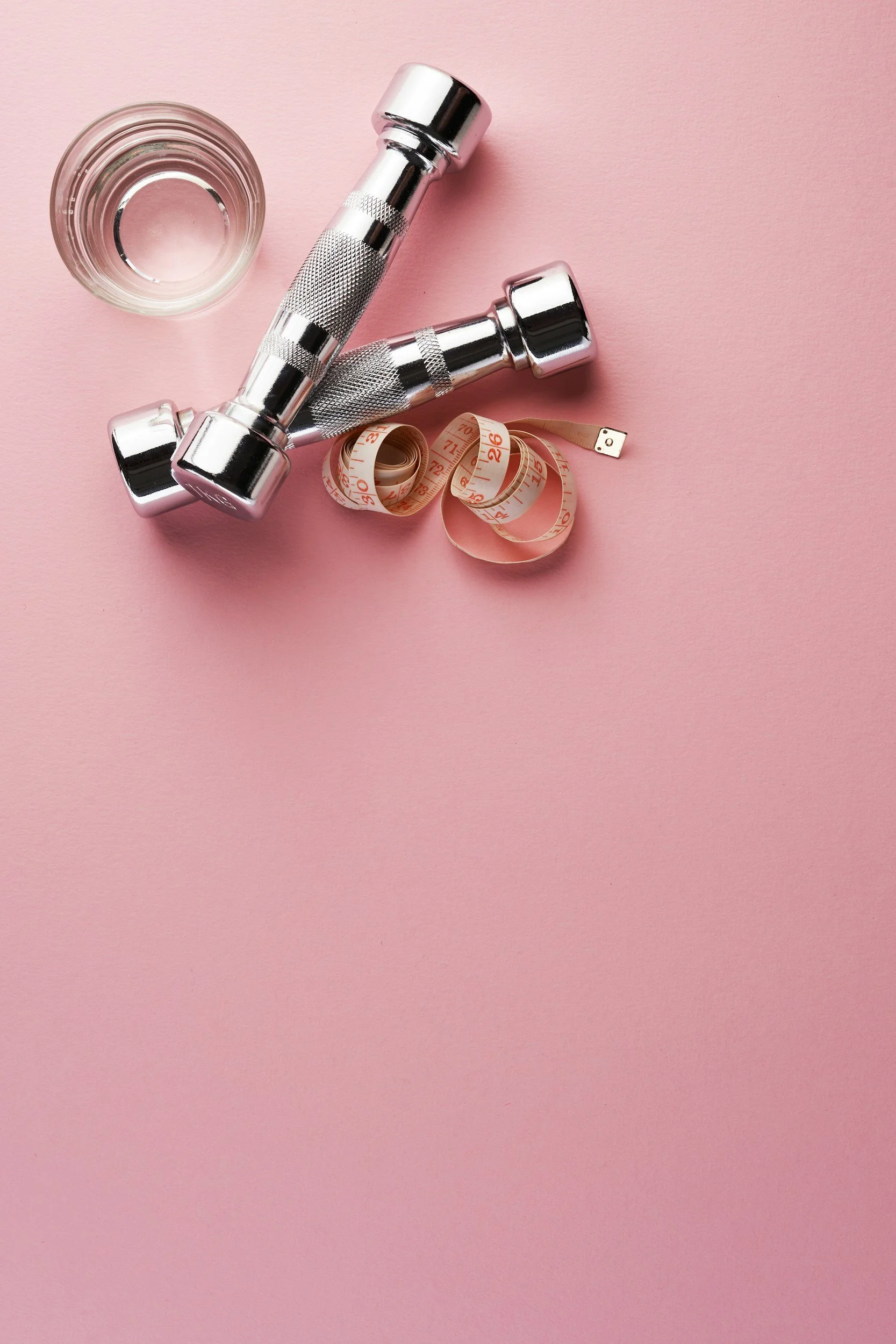 A glass of water, a pair of silver dumbbells, and a pink measuring tape on a pink background.