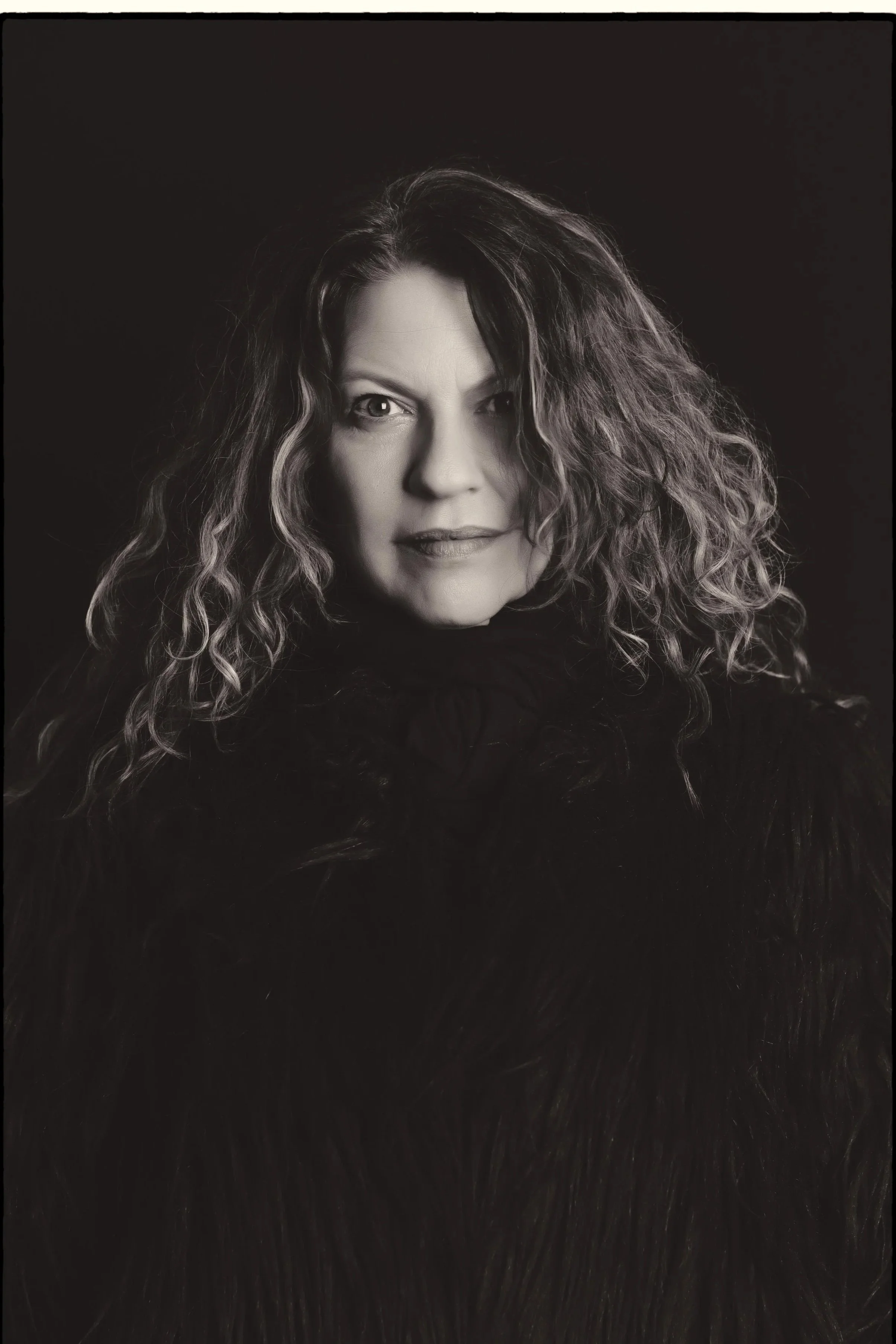 Black and white portrait of a spiritual holistic life coach philosopher healer  woman with curly hair, wearing a dark fur garment, looking directly at the camera.
