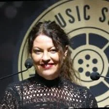 A woman smiling and speaking into two microphones at an event with a round banner in the background.