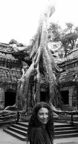 Witchy esoteric healer spiritual life coach Woman with long curly hair smiling in front of a large tree growing over ancient stone ruins.