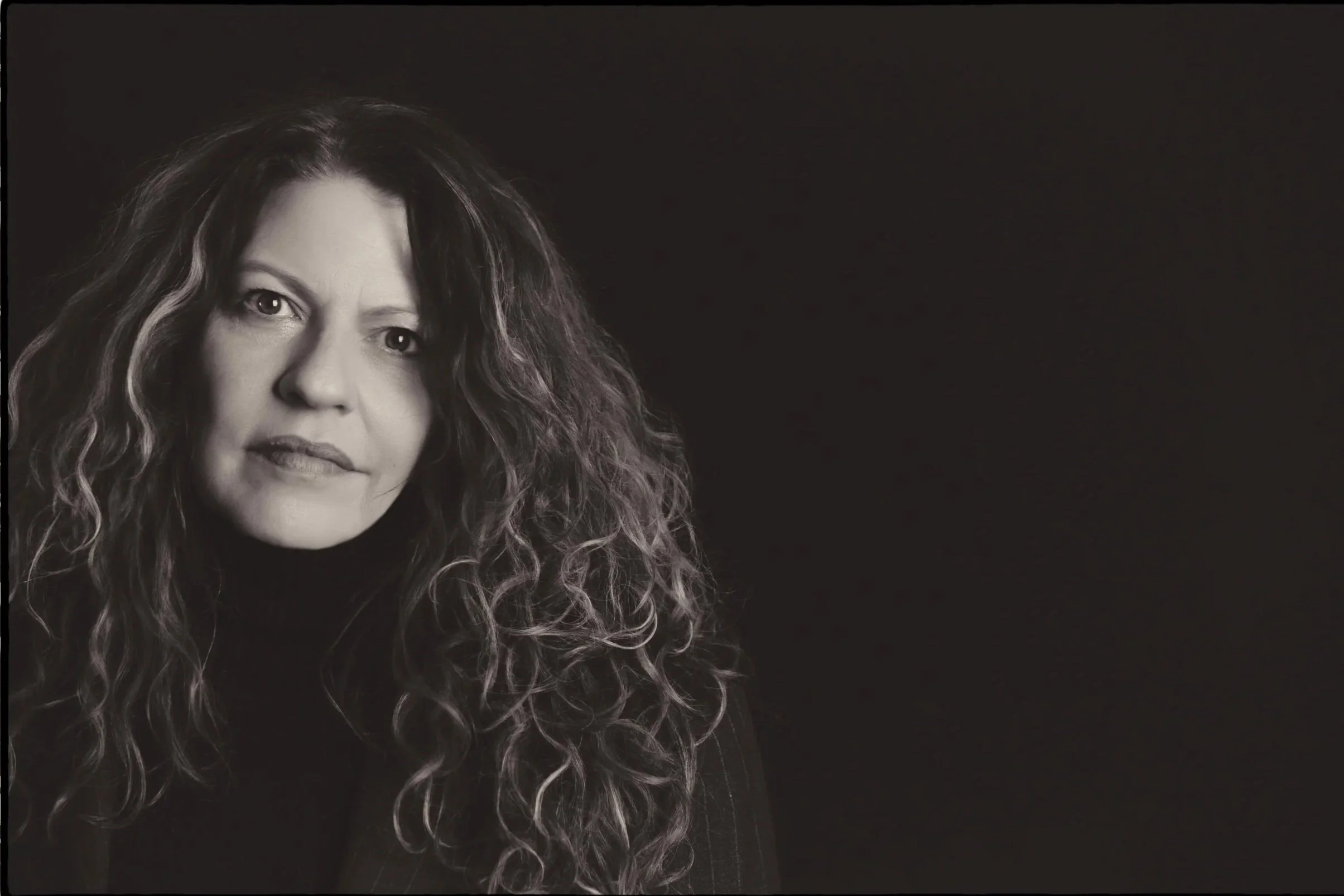 Black and white portrait of a spiritual holistic life coach philosopher healer woman with curly hair, wearing a turtleneck, looking at the camera with a neutral expression.