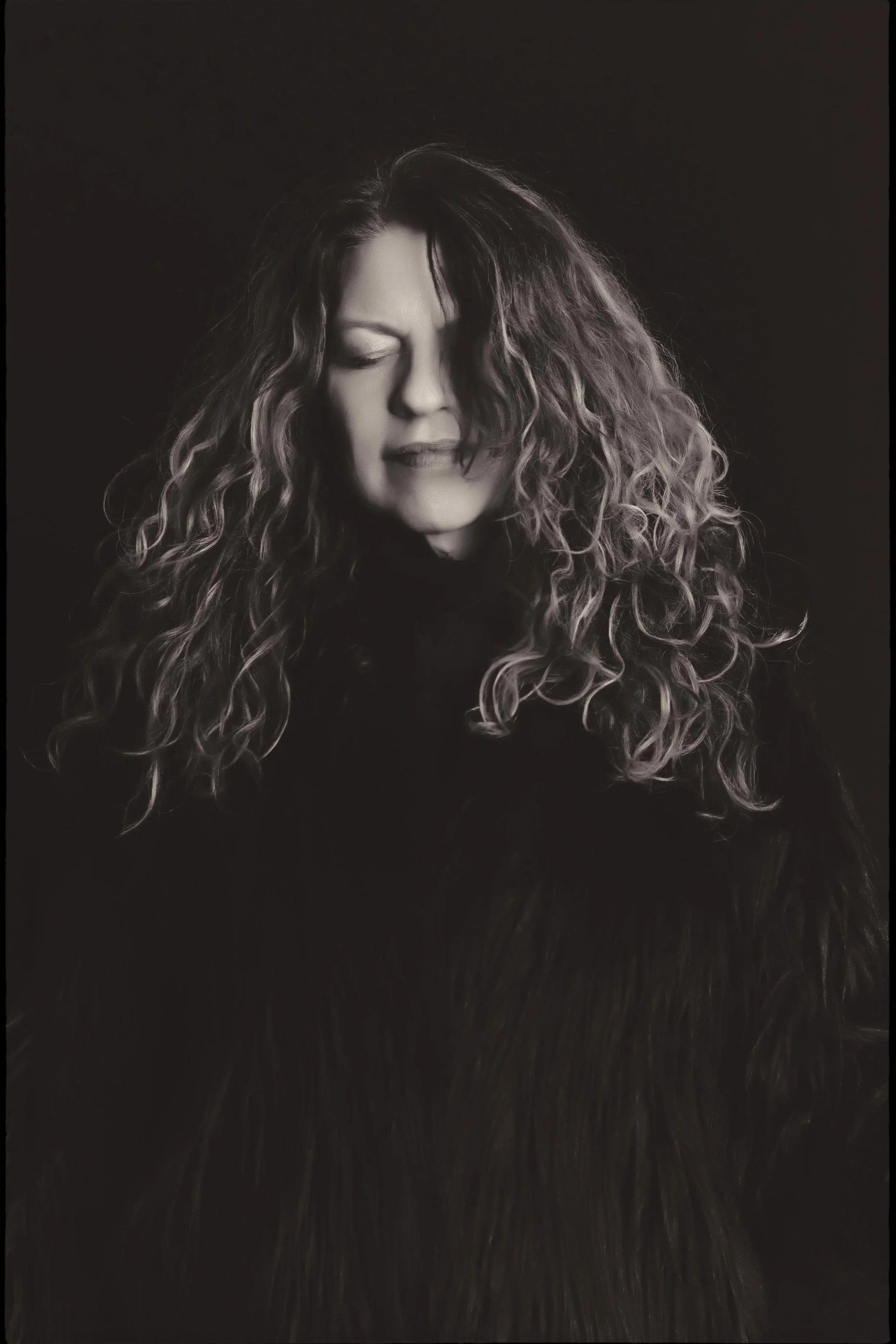 Black and white portrait of a spiritual holistic life coach philosopher healer  woman with curly hair, eyes closed, wearing a dark furry coat against a dark background.