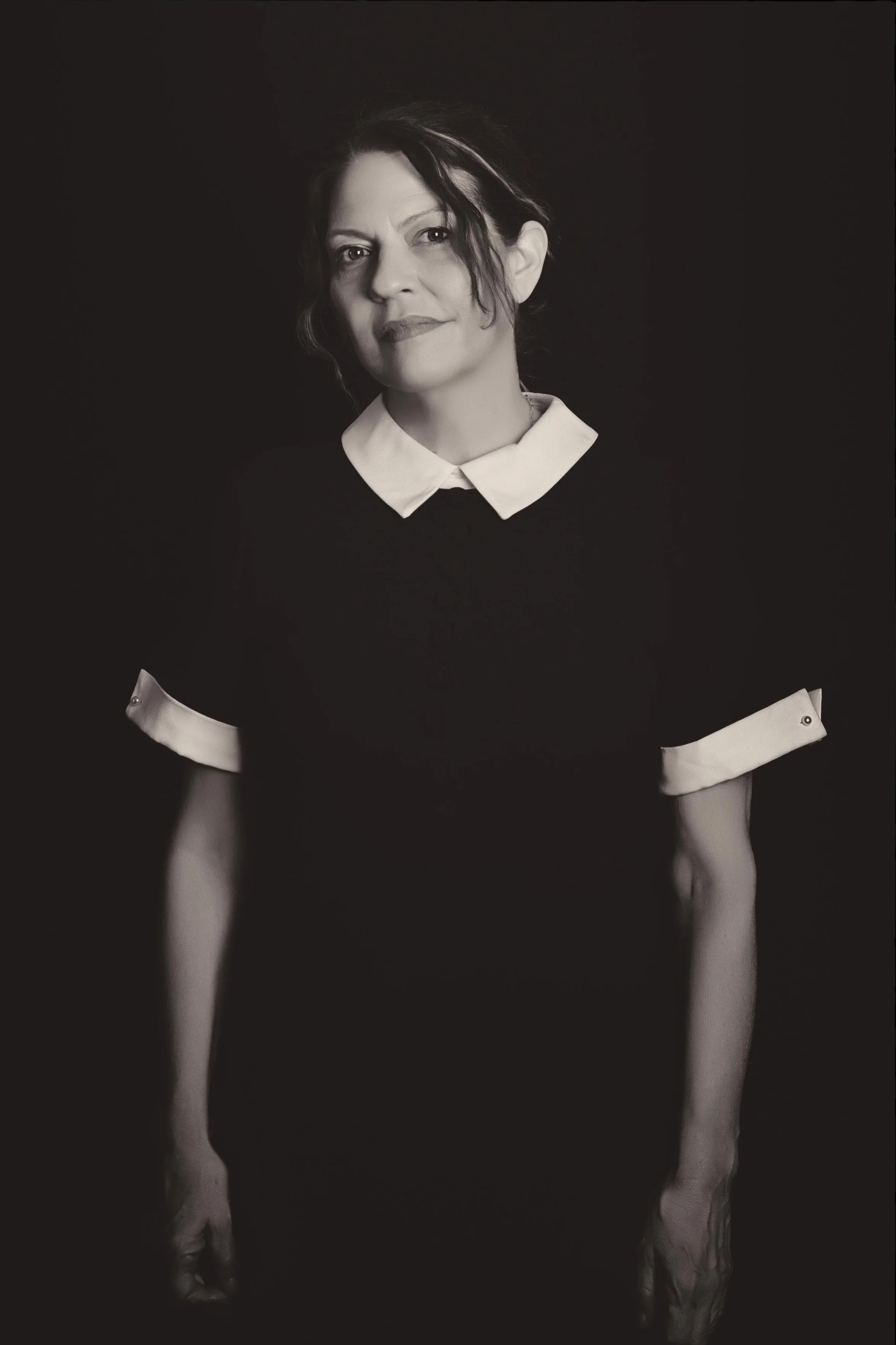 Black and white portrait of a spiritual holistic life coach philosopher healer  woman with short dark hair, wearing a dark dress with a white collar and cuffs, standing against a black background.