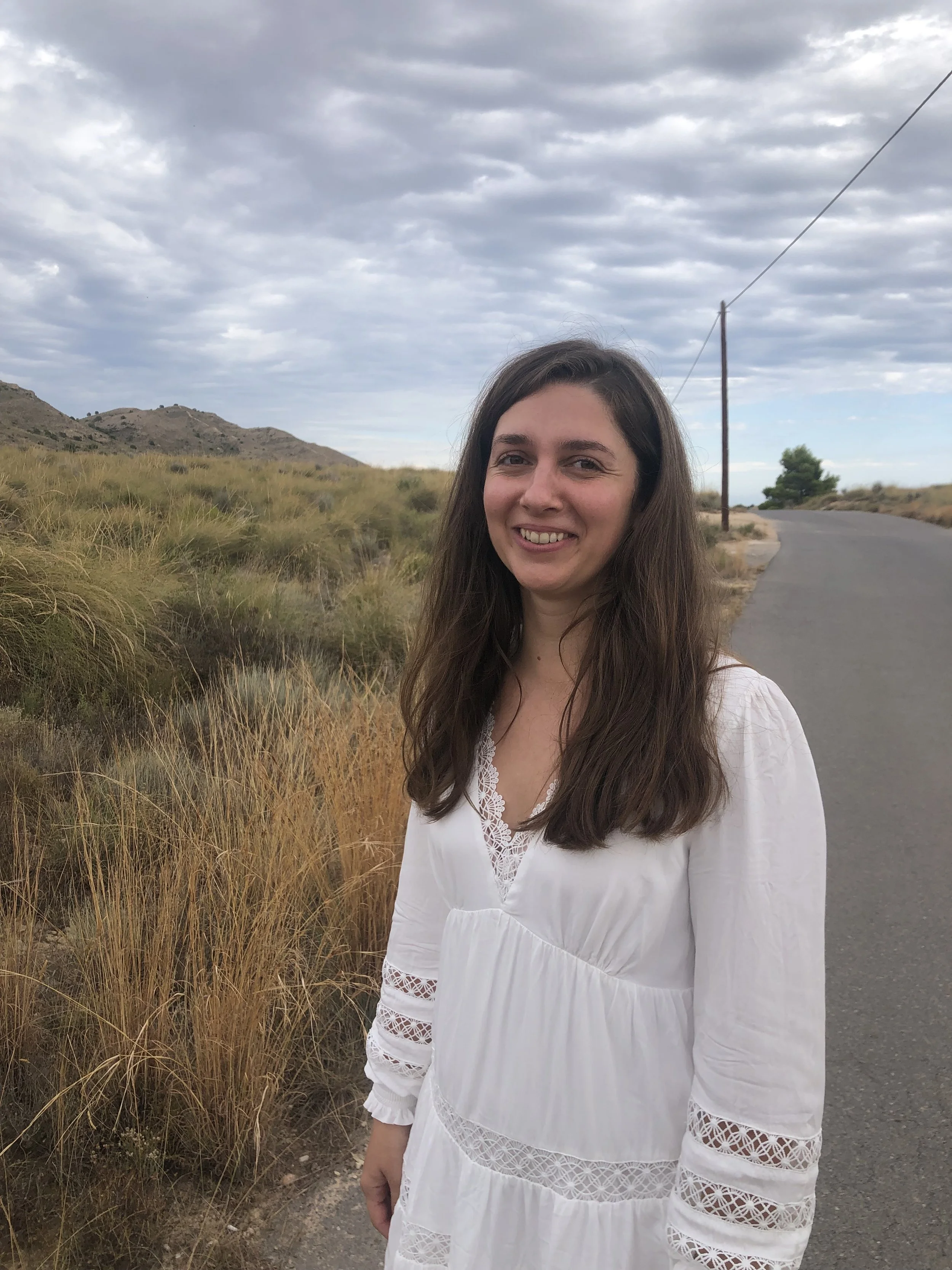 A young woman with long brown hair, smiling, wearing a white dress with lace details on the sleeves and neckline, standing on a rural road with grassy hills and a cloudy sky in the background.