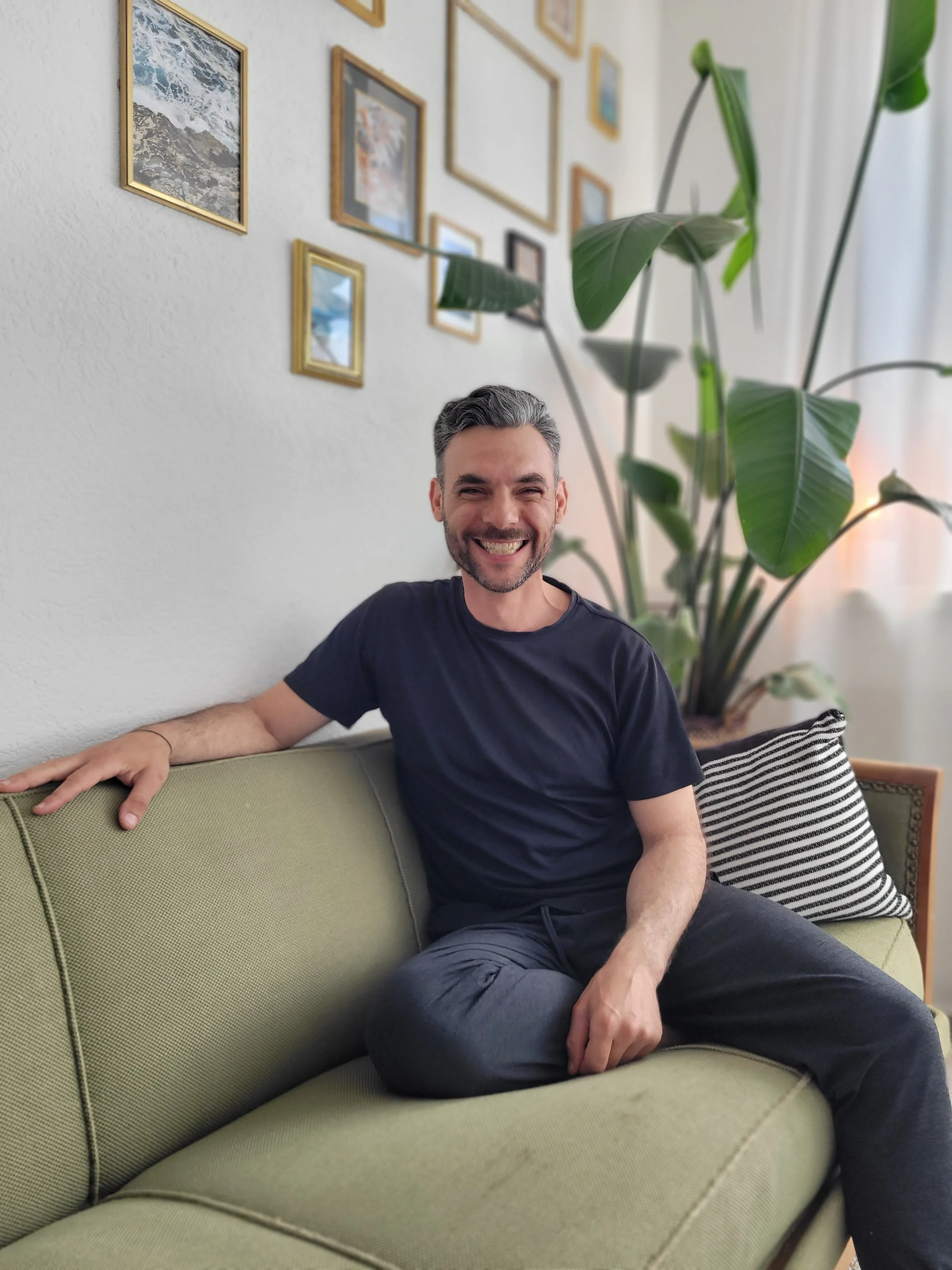 A smiling man with gray hair and beard sitting on a green couch in a living room, with a large potted plant and picture frames on the wall behind him.