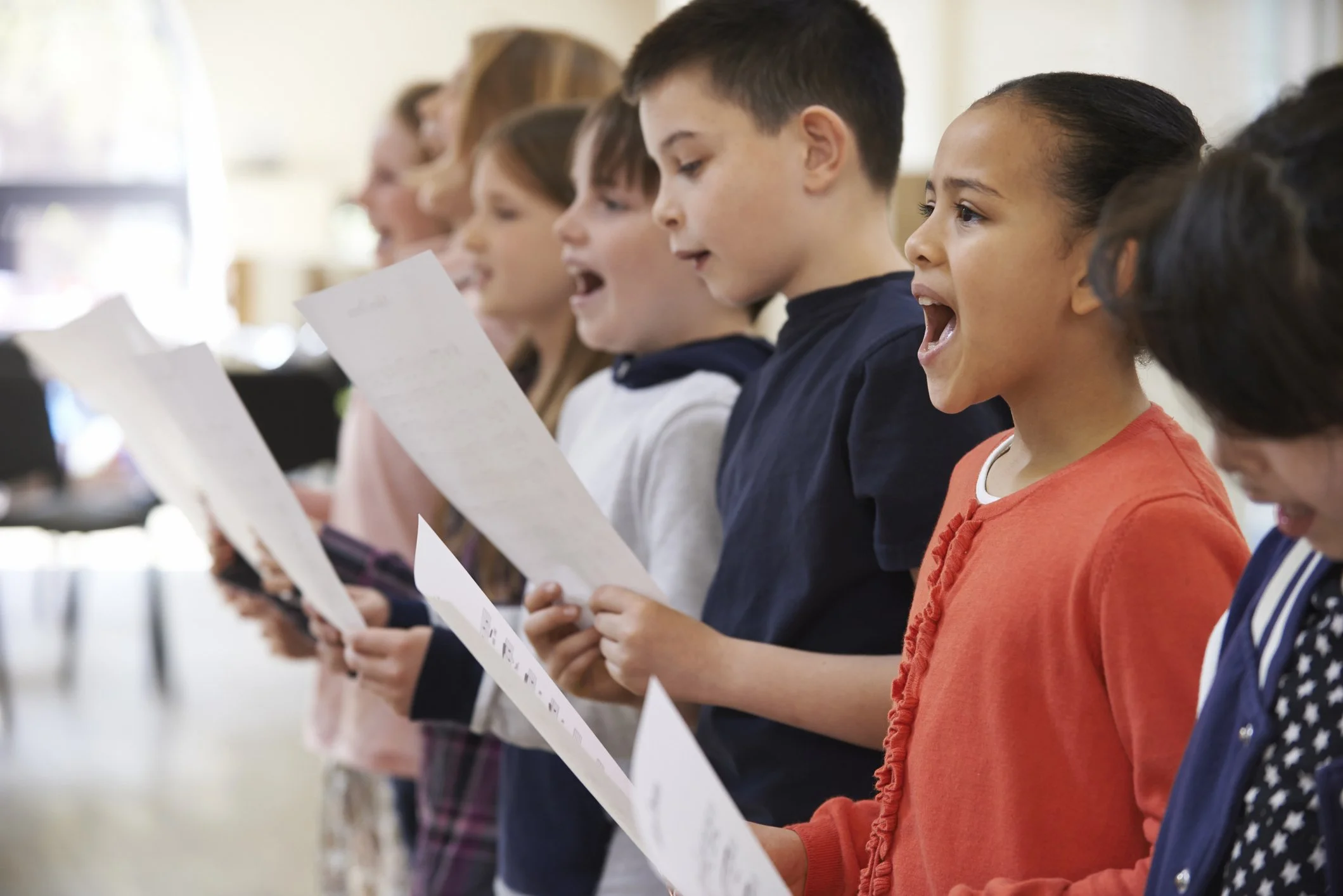 Children singing or reading aloud from sheets of paper in a classroom or assembly setting.