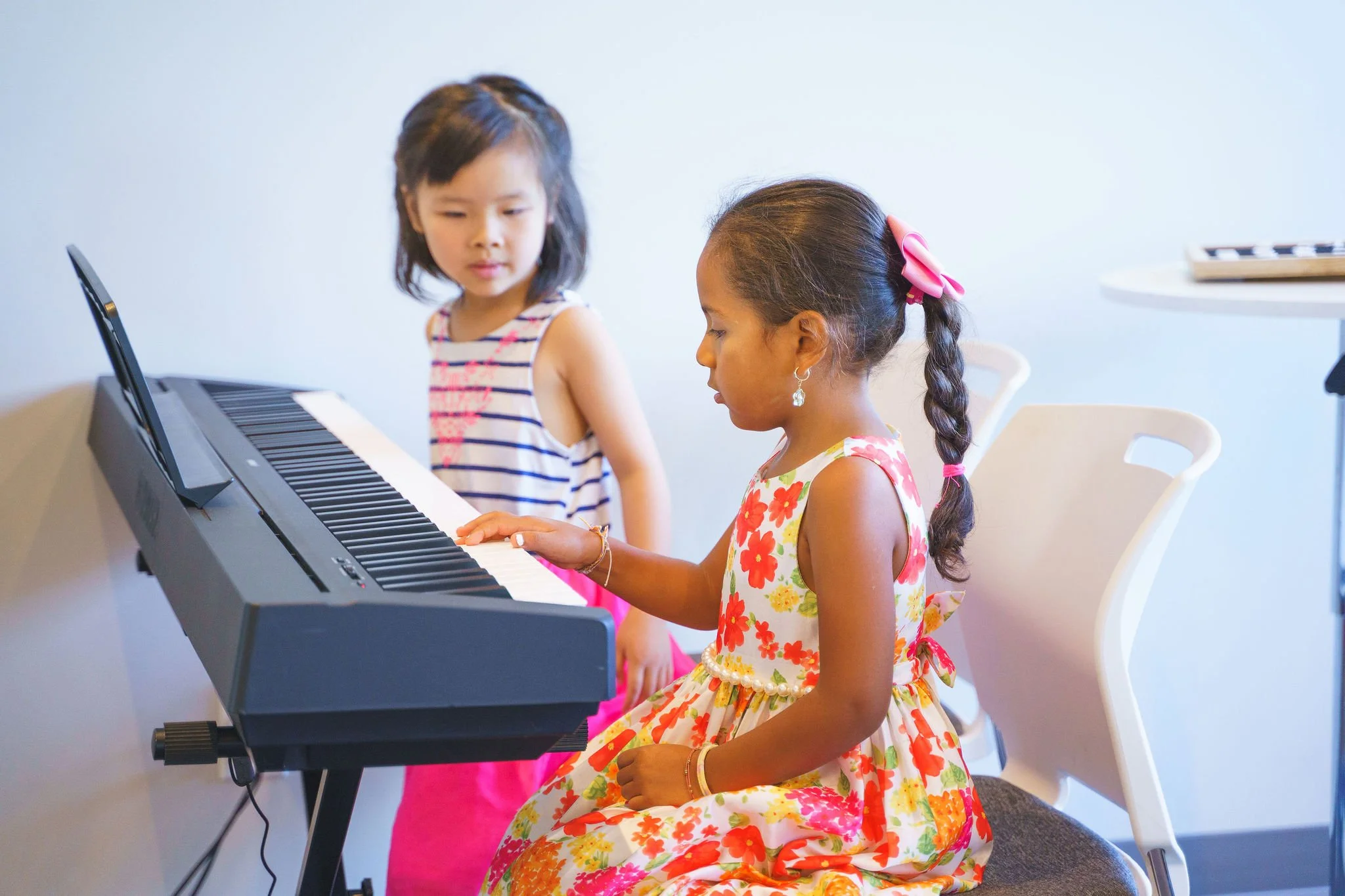 Two young girls, one sitting and playing the piano, the other standing beside her looking on, in a room with white walls.