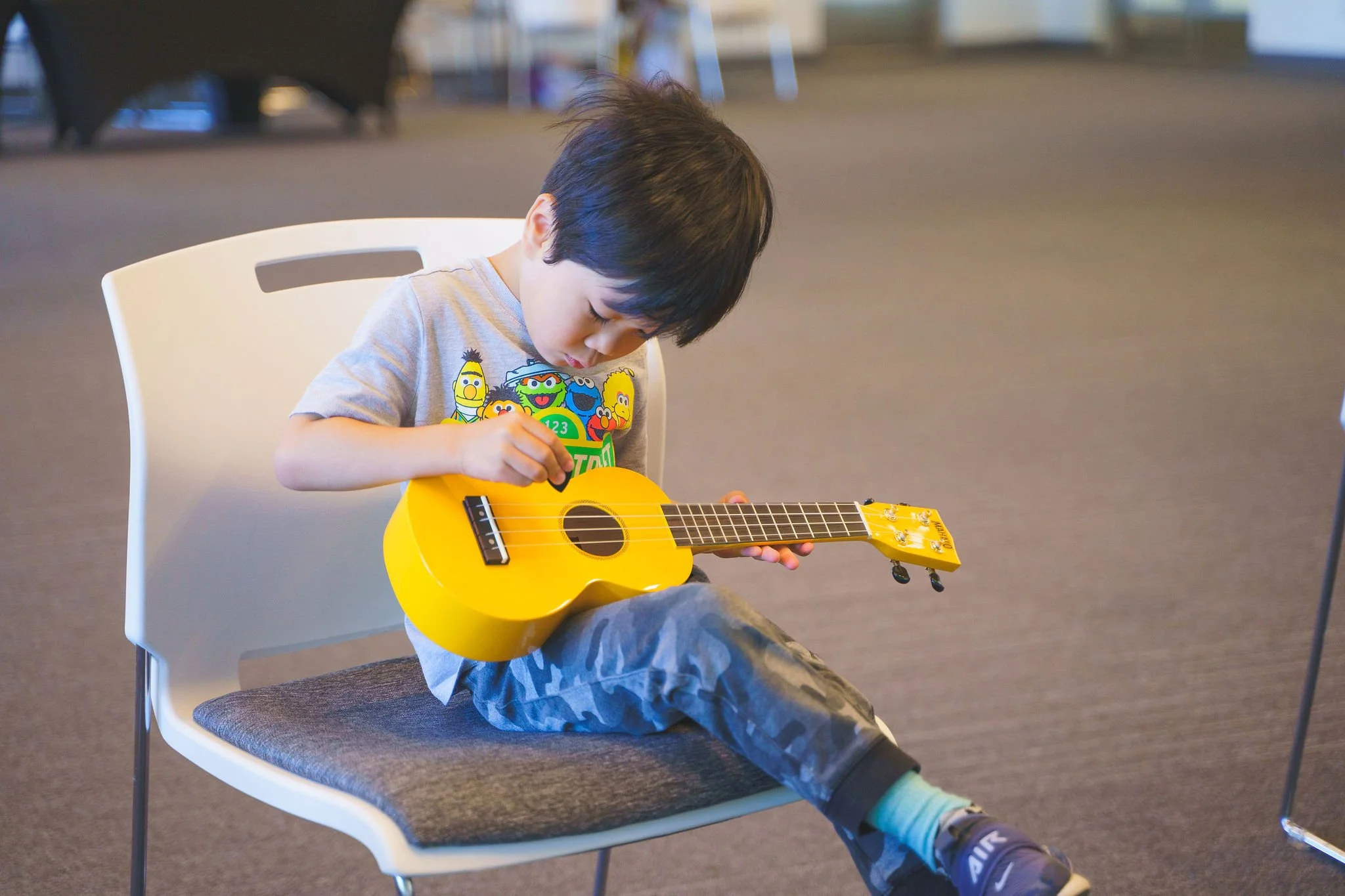 A young boy sitting on a chair playing a bright yellow ukulele.