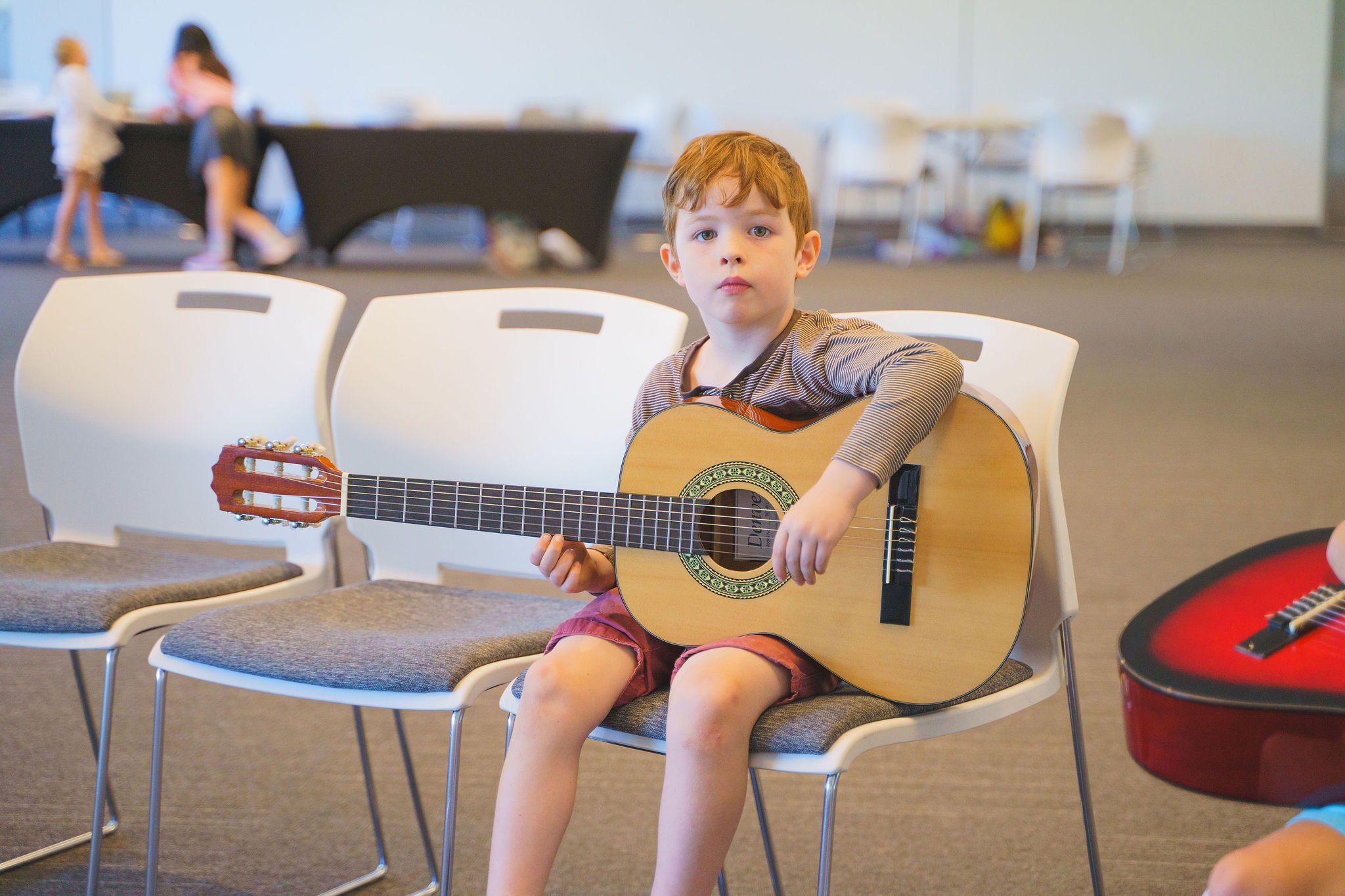 Young boy with red hair sitting on a white chair, holding an acoustic guitar, in a room with other children and chairs.