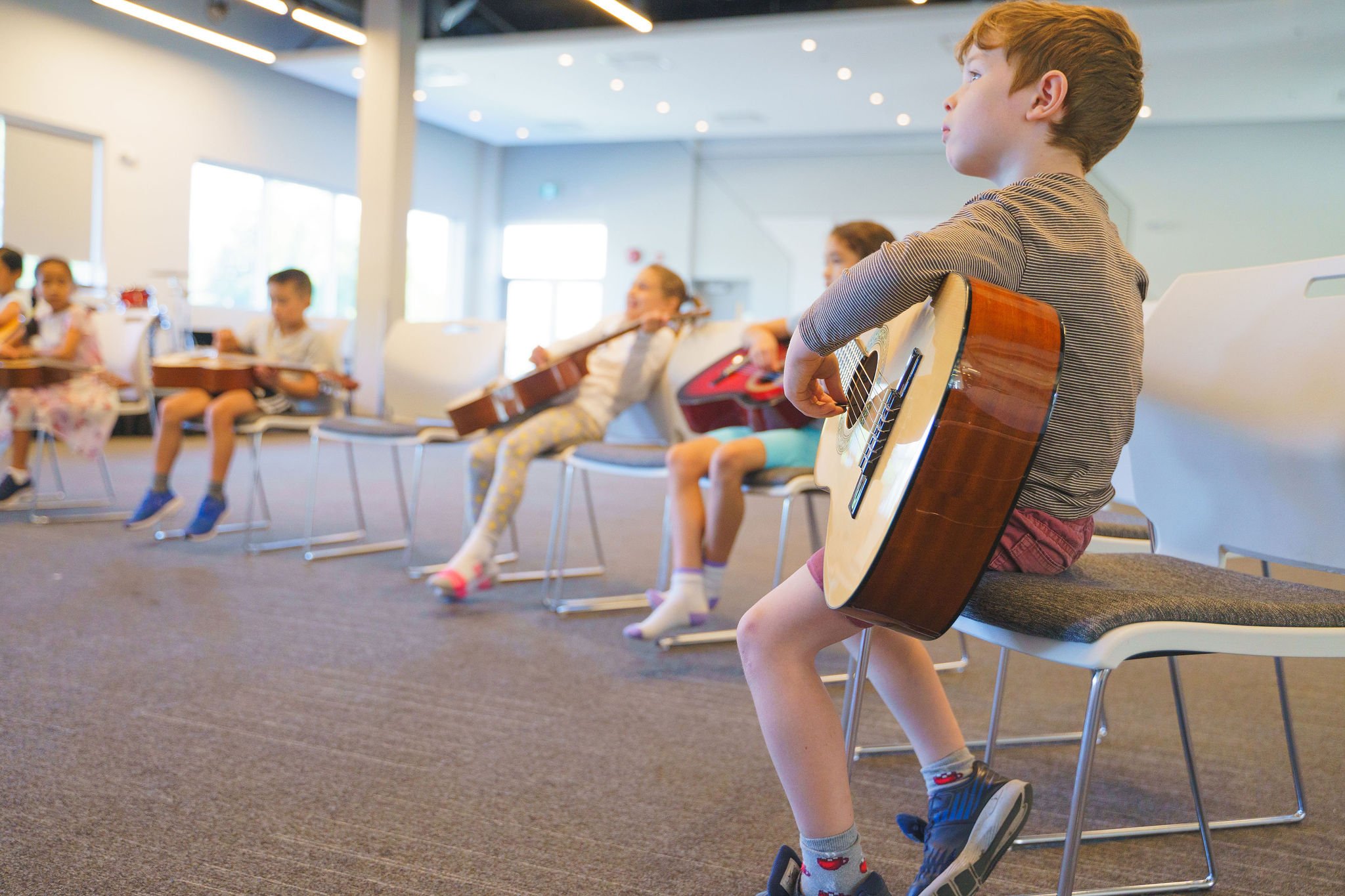 Children sitting on chairs in a music classroom, each holding or playing a guitar, with a focus on a young boy in the foreground playing an acoustic guitar.