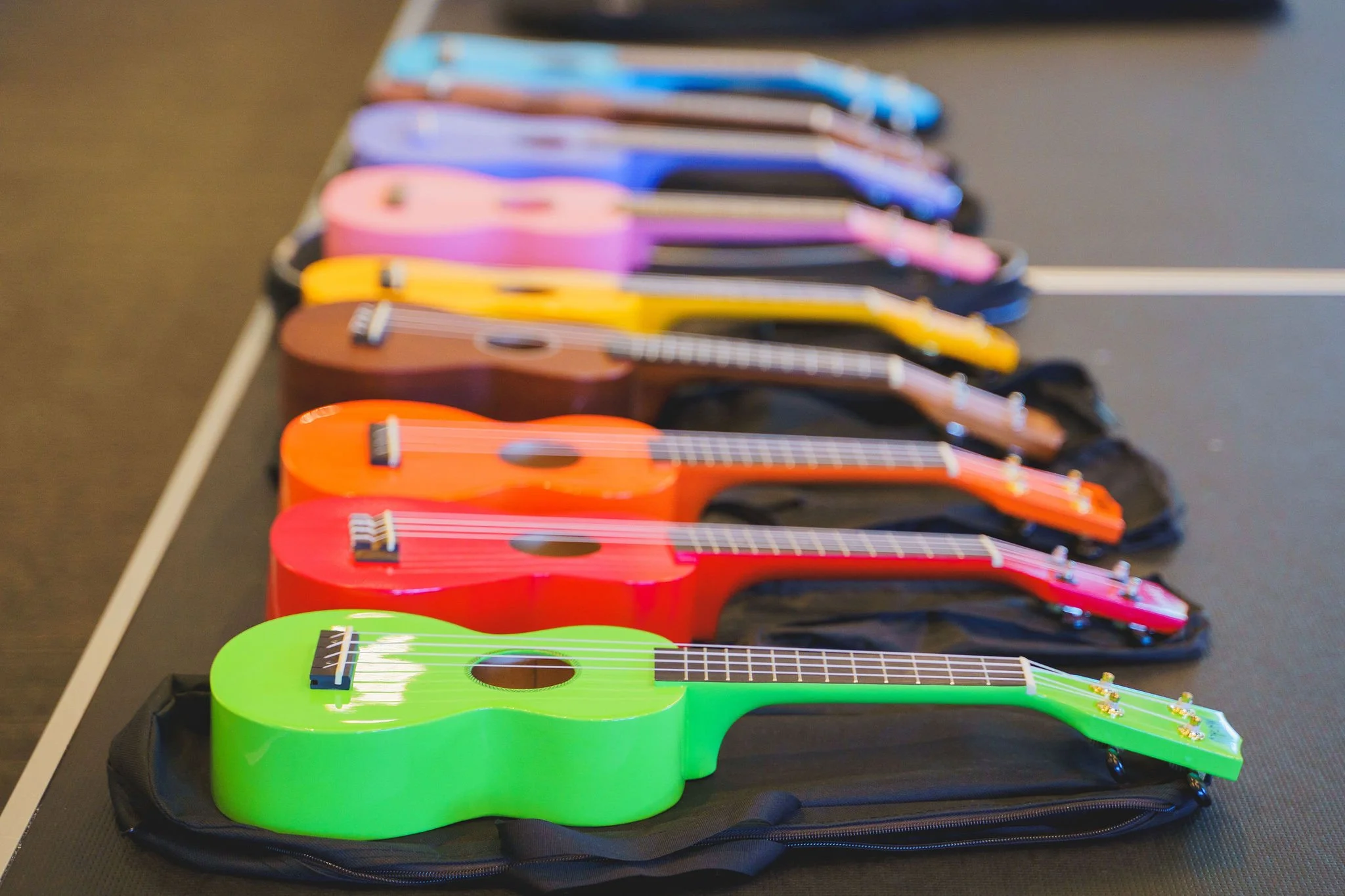 Colorful ukuleles in a row on black cases, arranged from green to pink, on a dark table.