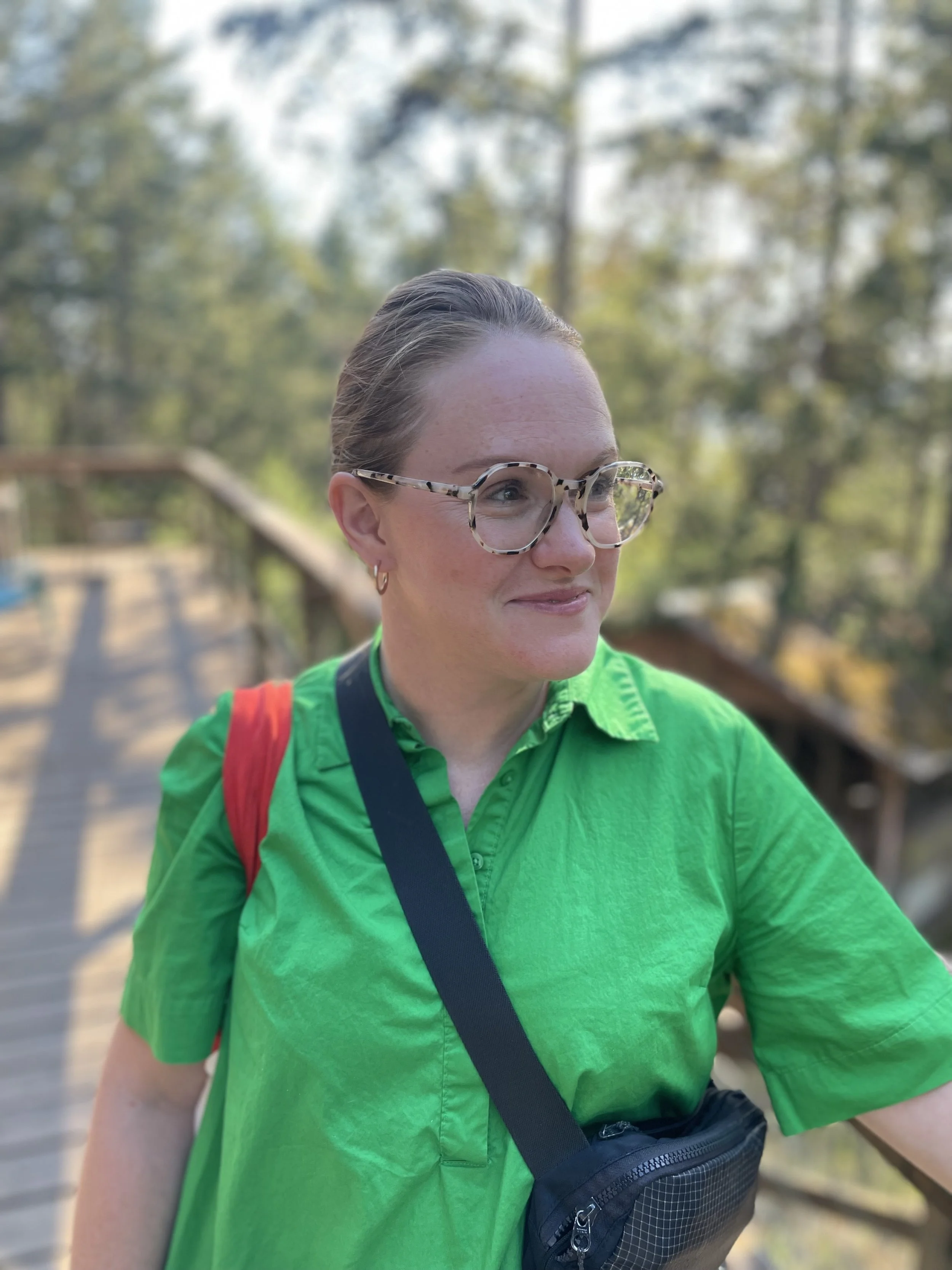 A woman with glasses and earrings wearing a green shirt and a red backpack, standing outdoors on a wooden bridge with trees in the background.