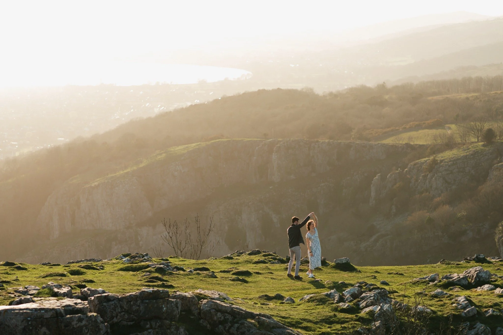 A couple dancing outdoors in a grassy area with rocks, hills, and a body of water in the background during sunset.