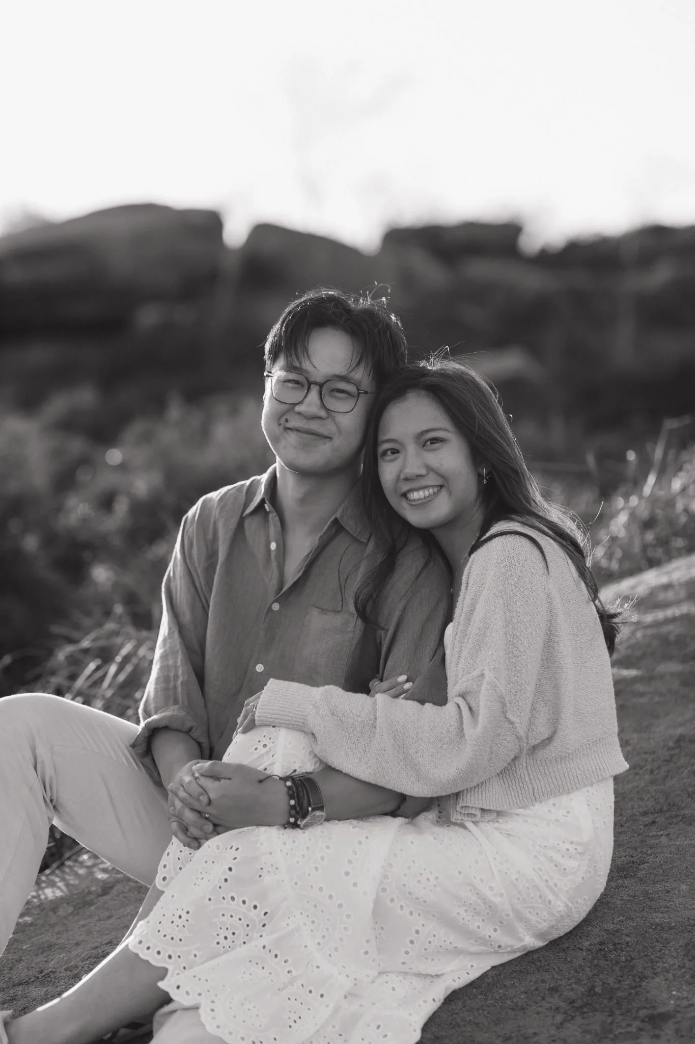 A smiling young man with glasses and a woman with long dark hair sitting close together outdoors, with a rocky landscape and a tree in the background, both looking happy.
