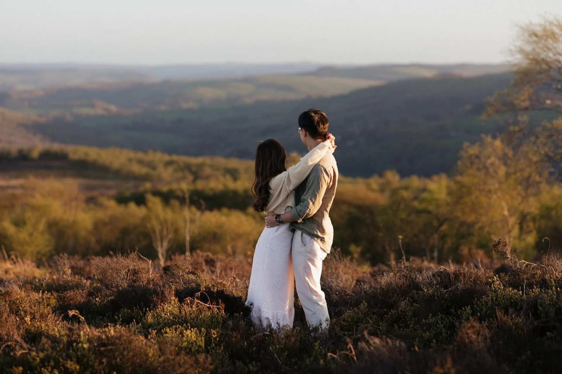 A couple standing in a field at sunset, embracing each other with hills in the background.